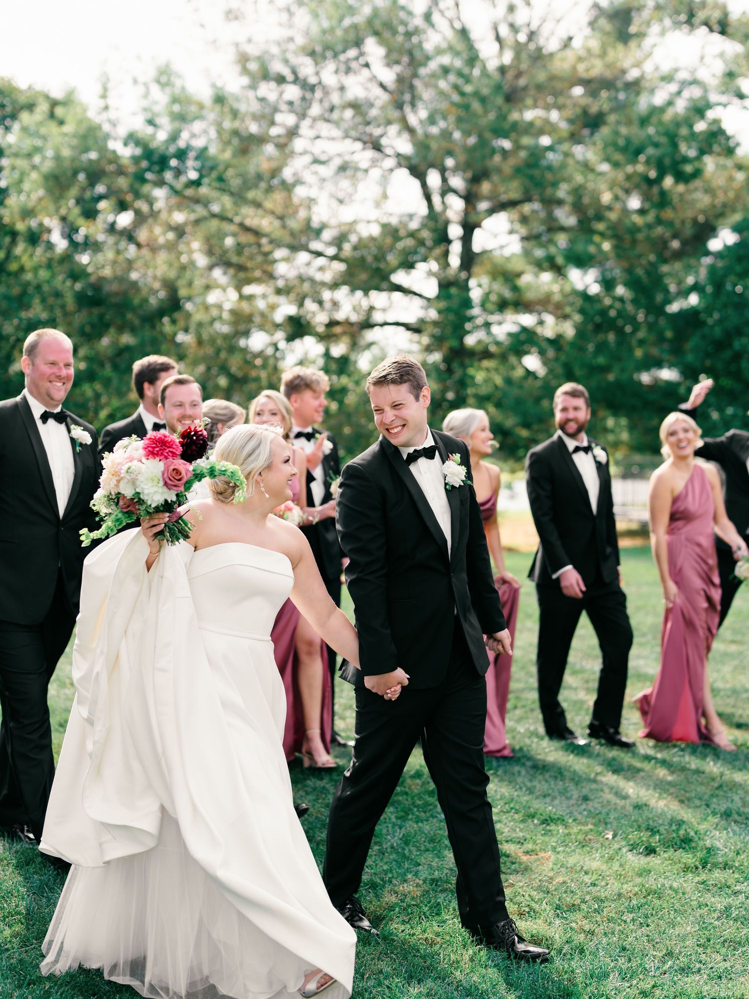 Wedding party in formal attire walks together on grassy lawn during outdoor celebration.