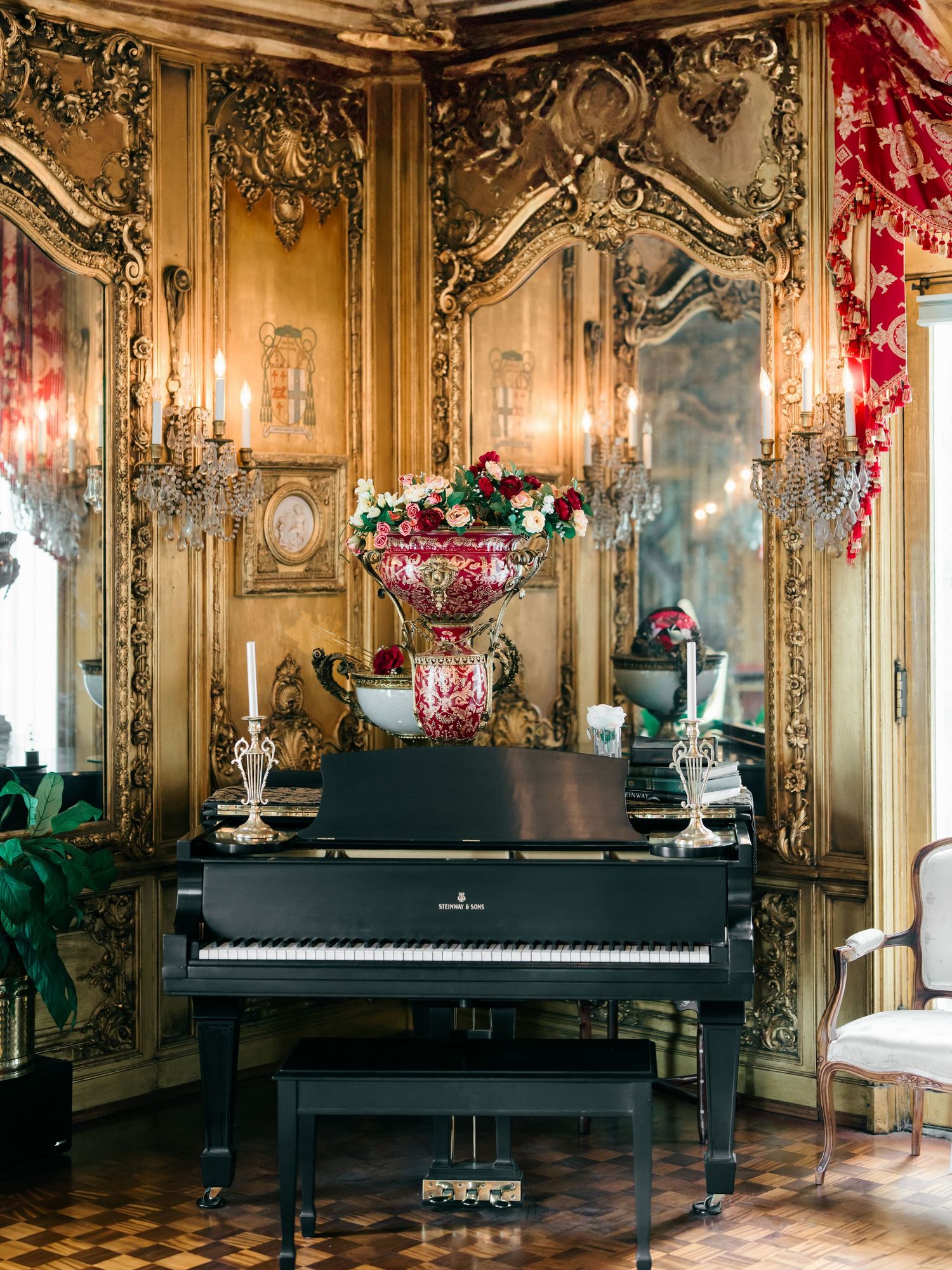 A grand piano sits in an ornate baroque room with gilded walls, crystal chandeliers and elegant red drapery.