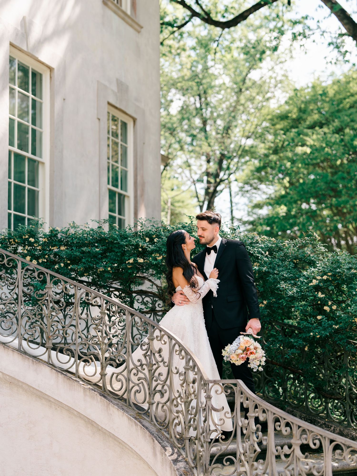 Bride and groom share a romantic moment on an ornate iron staircase outside an elegant estate surrounded by lush greenery.