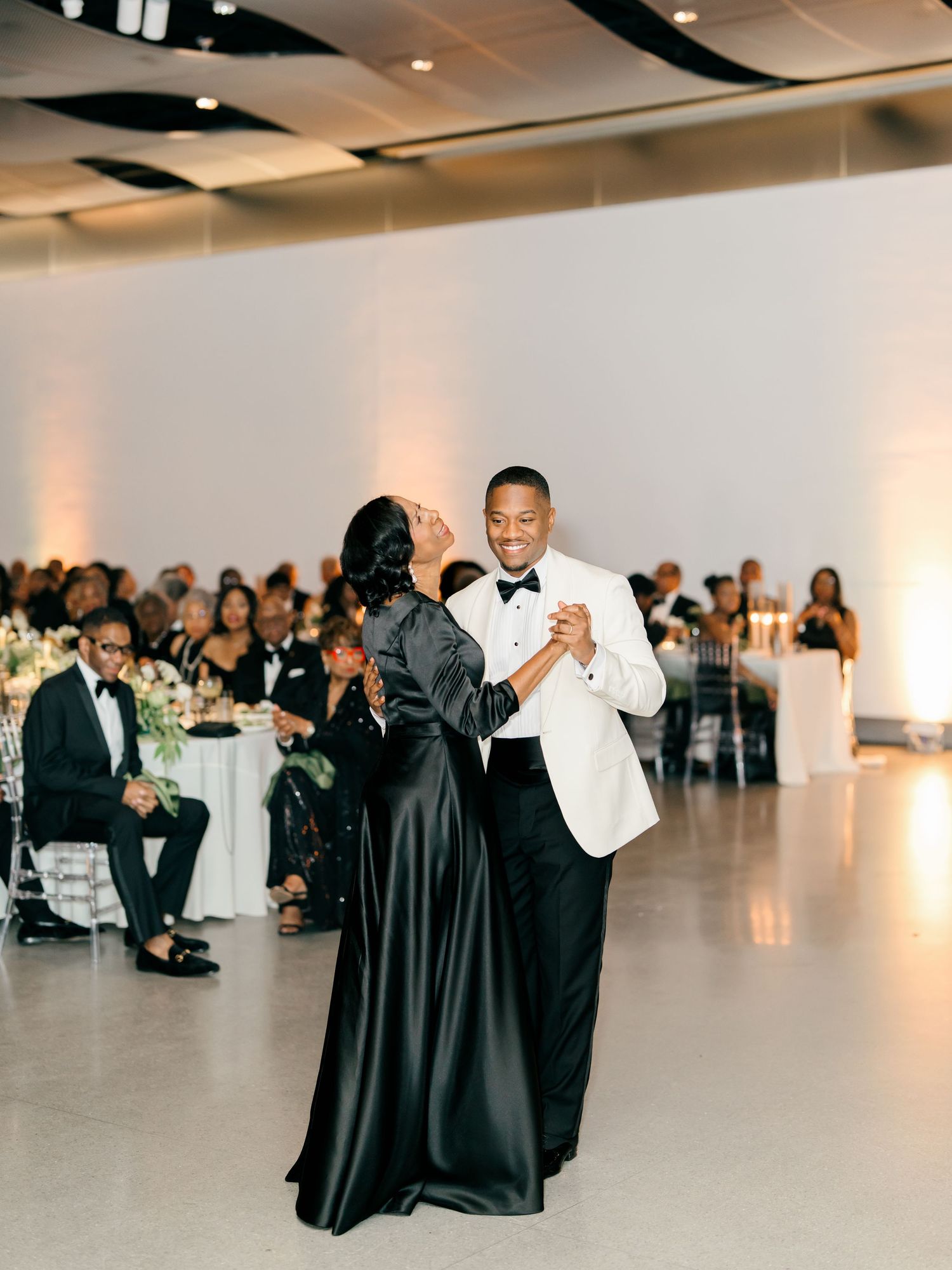A couple shares an elegant dance at their wedding reception in a modern white venue with guests looking on.