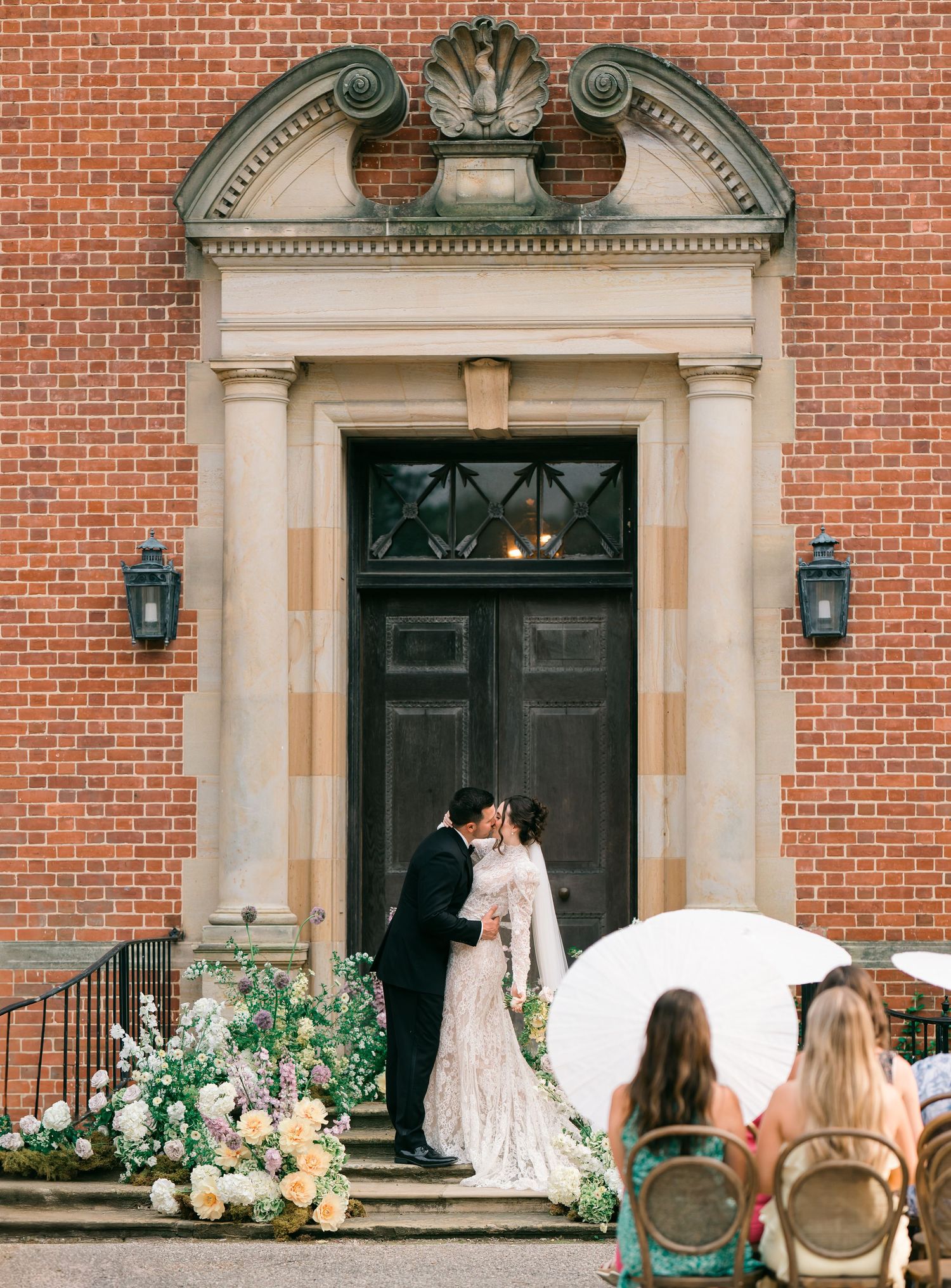 Elegant couple shares a romantic moment at brick building entrance adorned with floral arrangements and white umbrella.