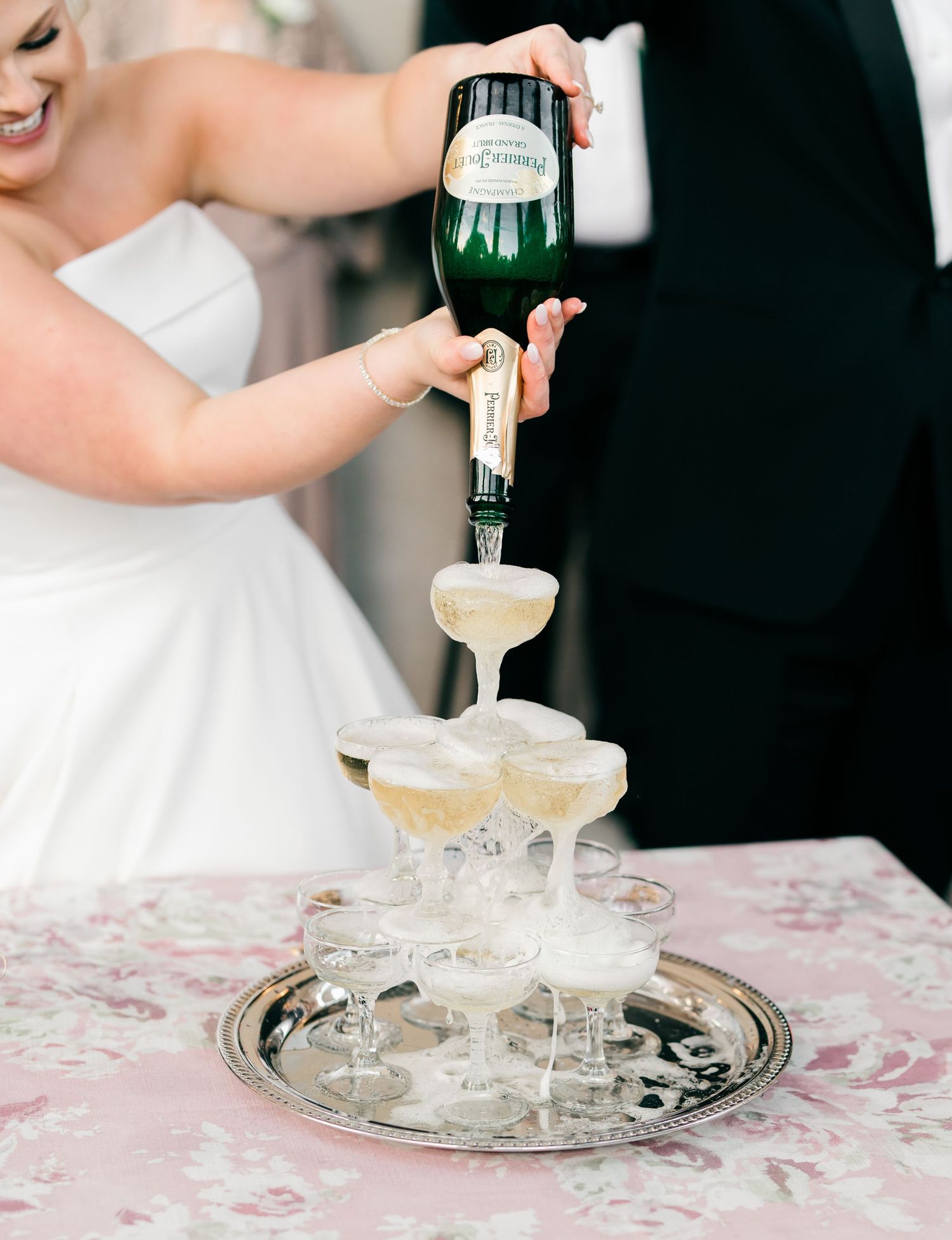 Champagne being poured into a tower of coupe glasses arranged on a silver tray with a floral tablecloth.