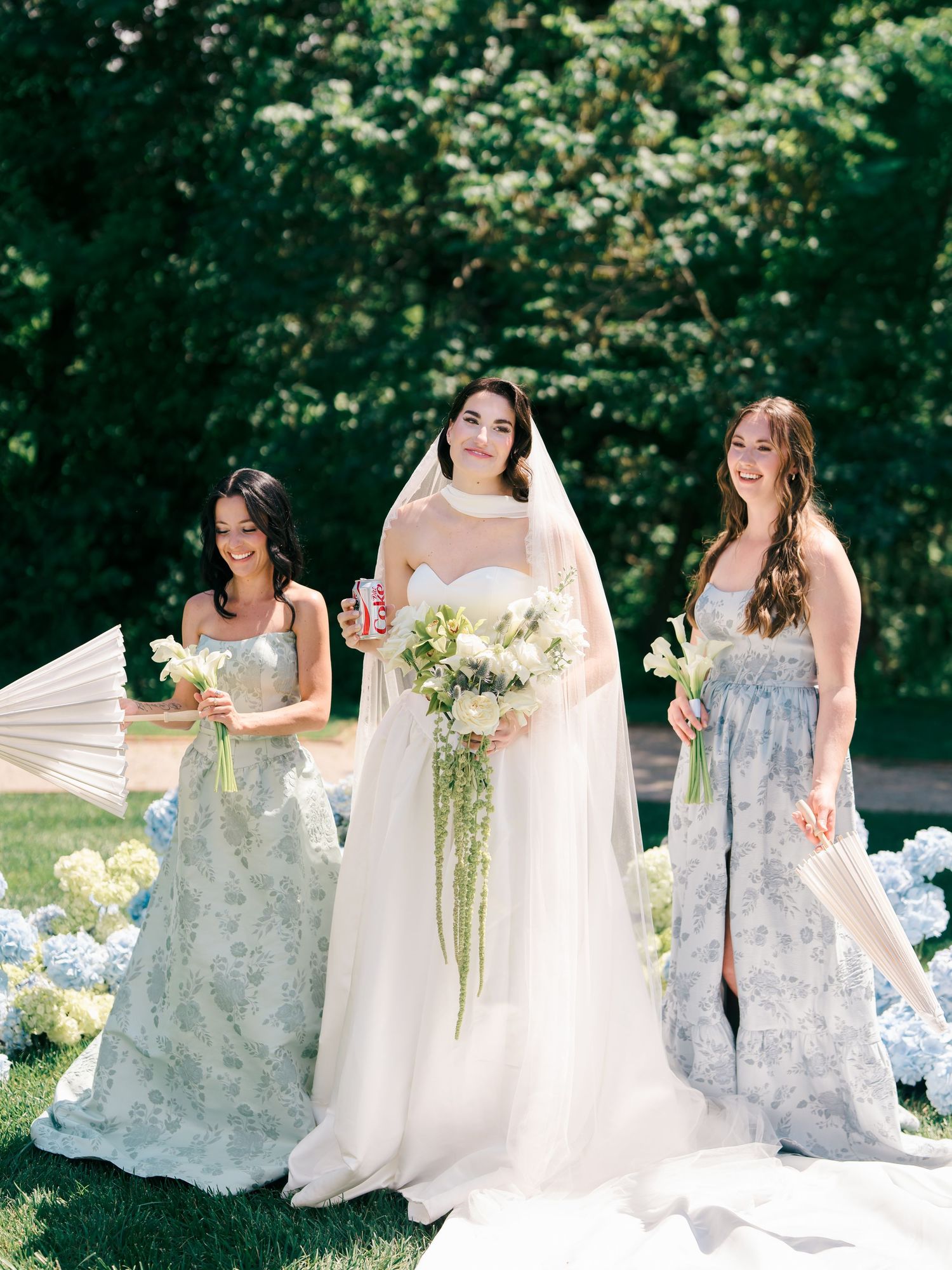 Bride in white gown with veil holds floral bouquet, flanked by two bridesmaids in floral dresses outdoors.
