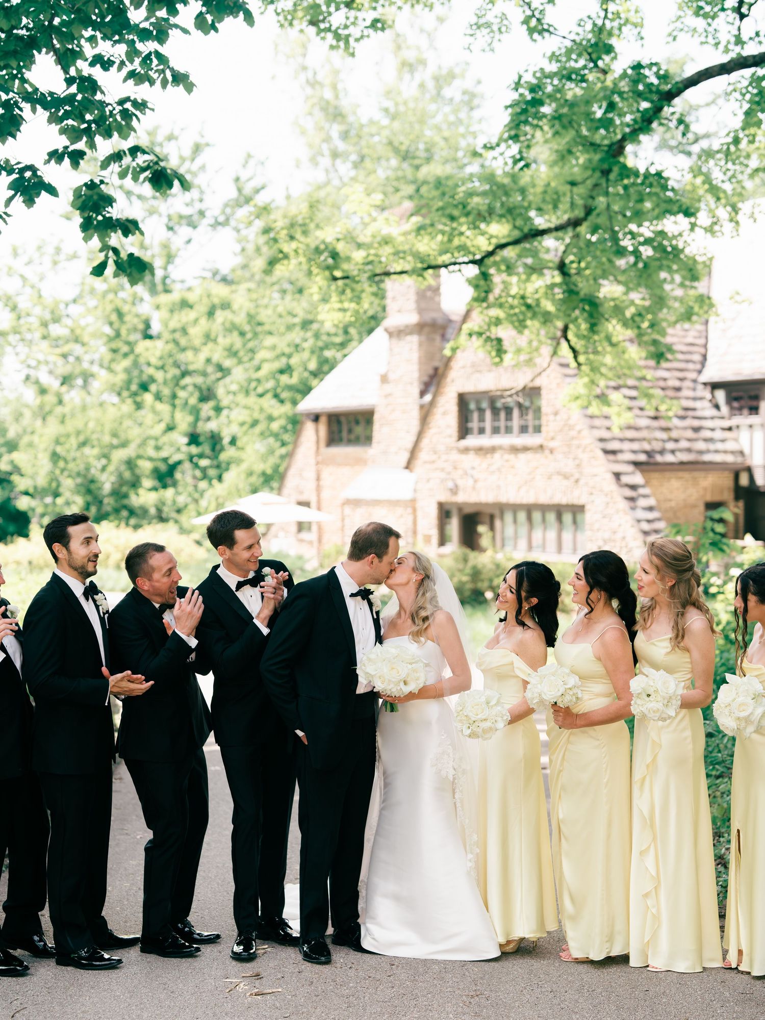 Wedding party poses outdoors, bride and groom kissing, groomsmen in tuxedos, bridesmaids in yellow gowns, historic building backdrop. Ohio.
