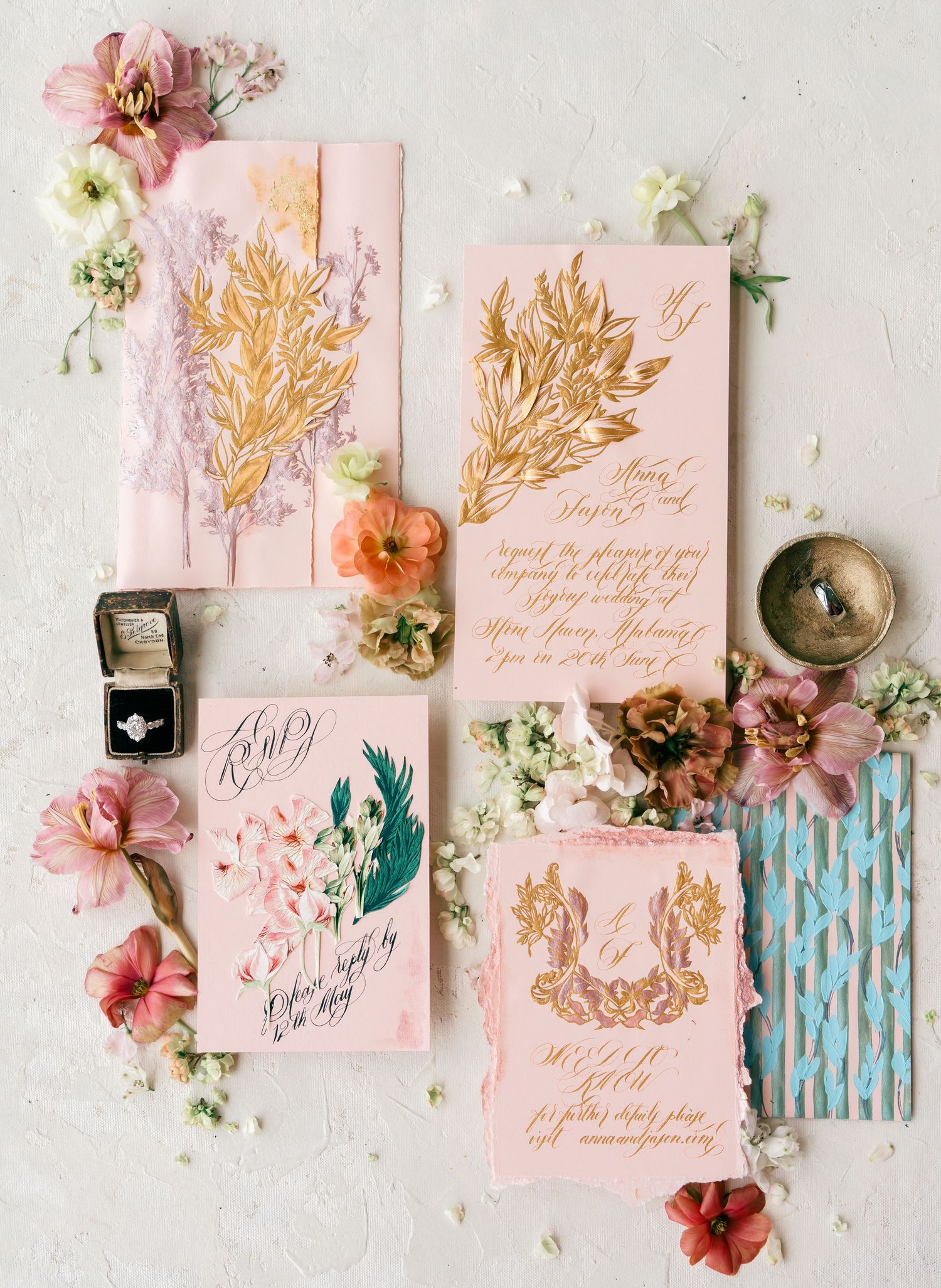 Elegant wedding stationery suite with gold botanical designs on blush pink cards, surrounded by colorful flowers and a diamond ring.