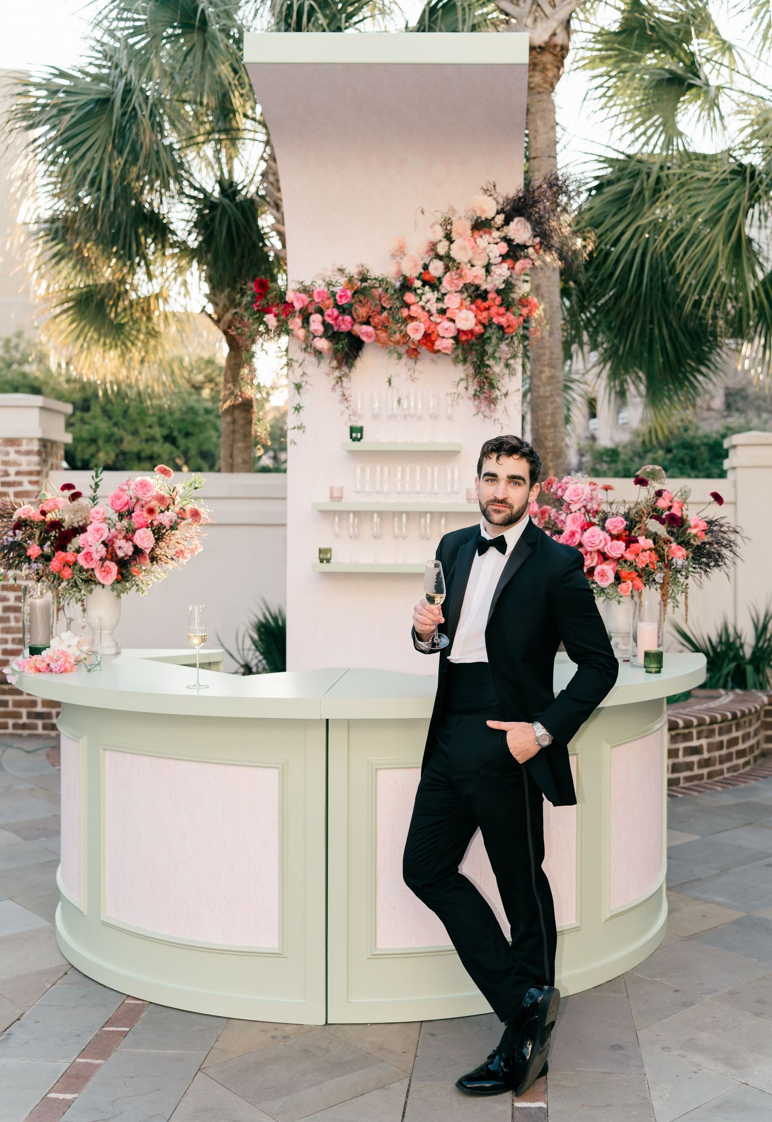 White curved bar decorated with pink floral arrangements and palm trees in an outdoor setting.