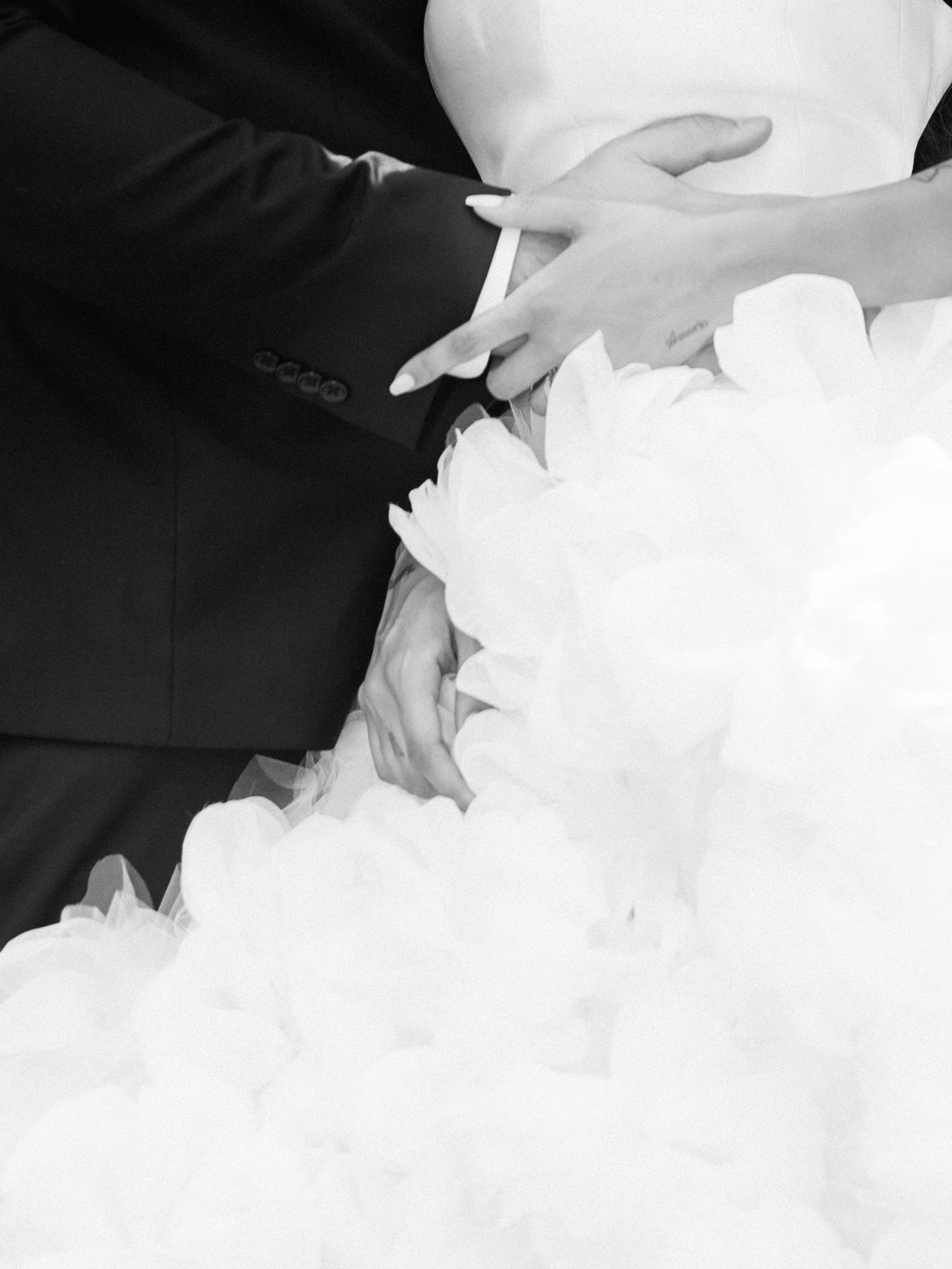 Black and white wedding photo of bride and groom holding hands, showcasing elegant white ruffled bridal gown.
