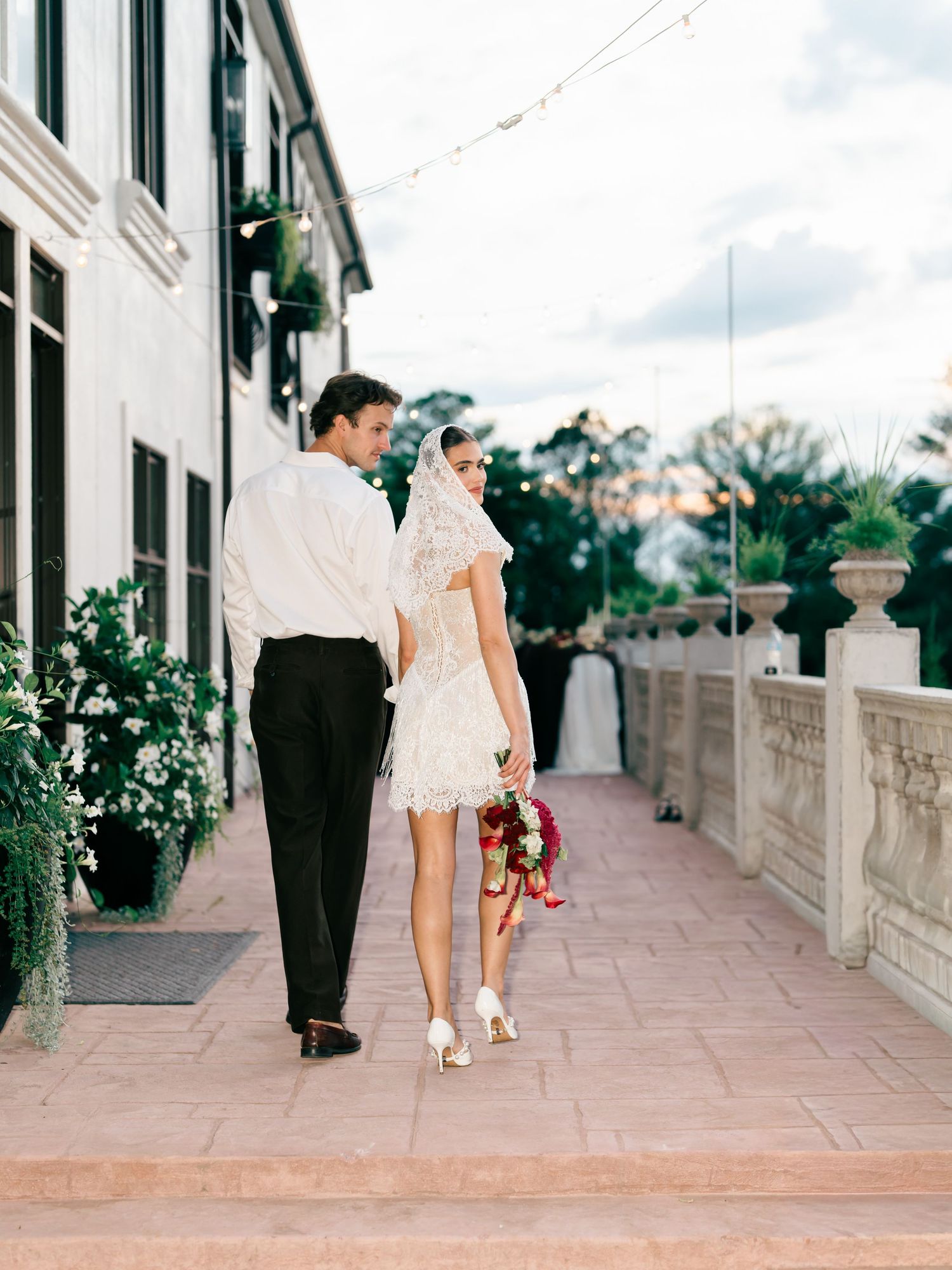 Bride in lace mini dress and veil walks with groom in black trousers along elegant terrace, holding red bouquet.