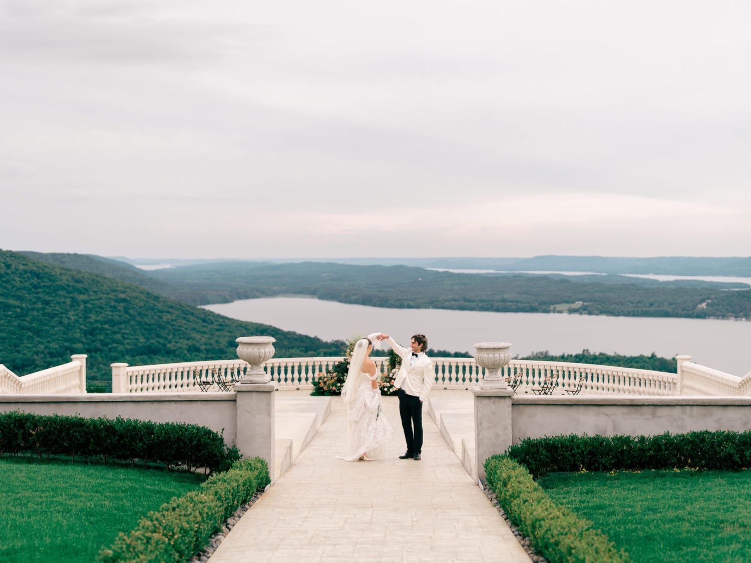 Bride and groom share romantic moment on elegant terrace overlooking scenic river and lush green hills.