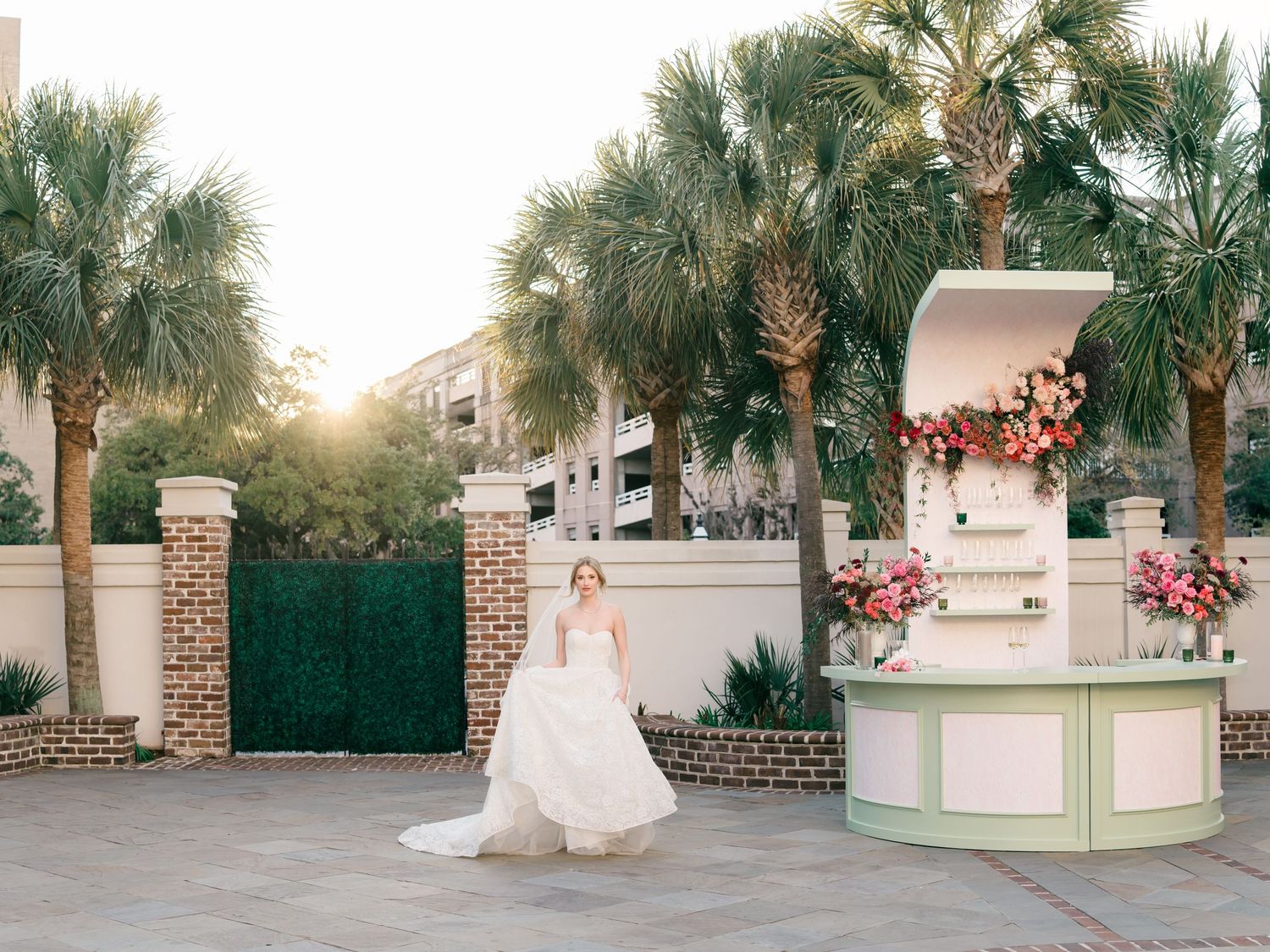 A bride in a white wedding gown stands gracefully in front of palm trees and brick columns during golden hour.