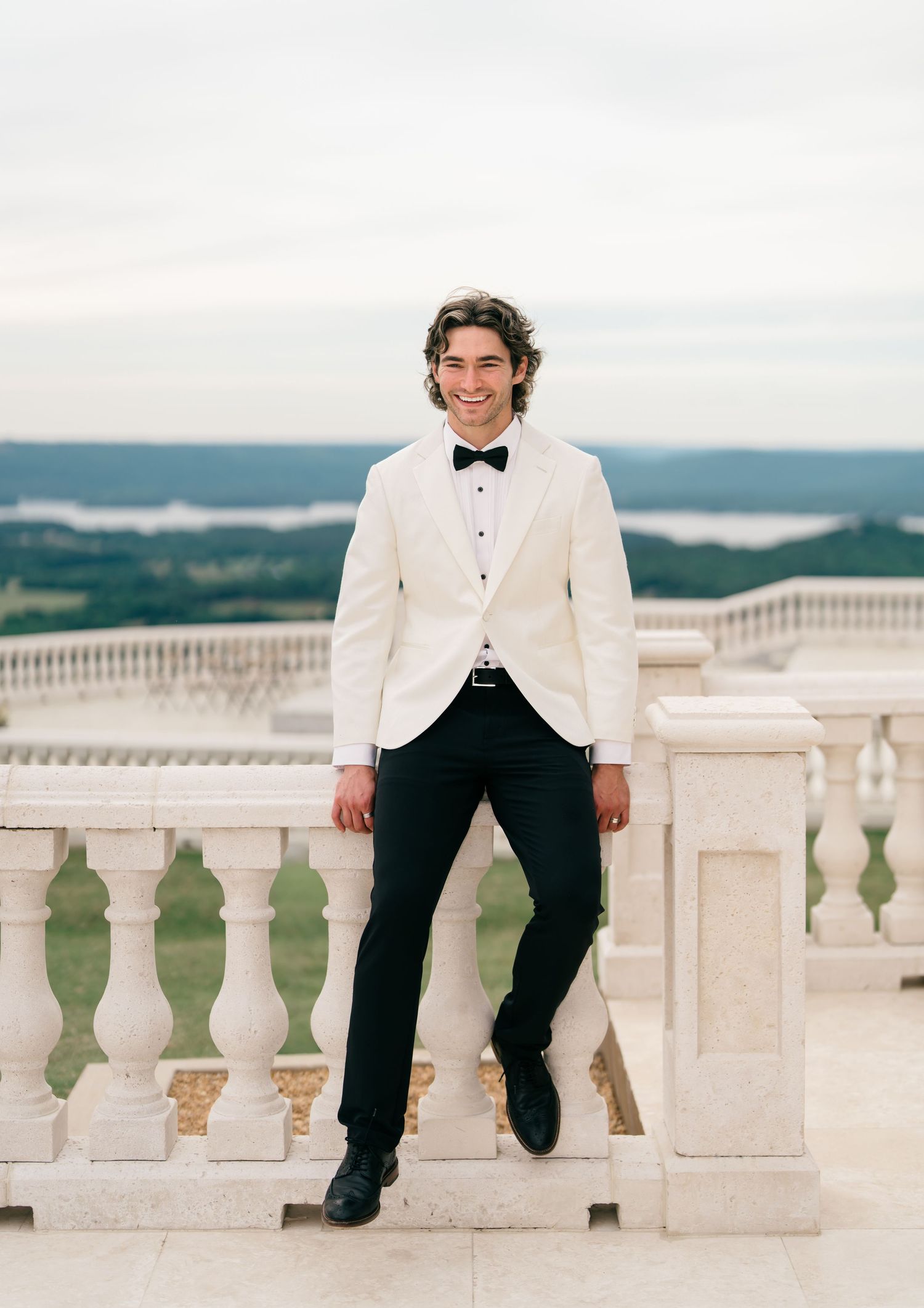 Smiling groom in white tuxedo jacket and black bow tie leaning on stone balustrade with scenic landscape view.