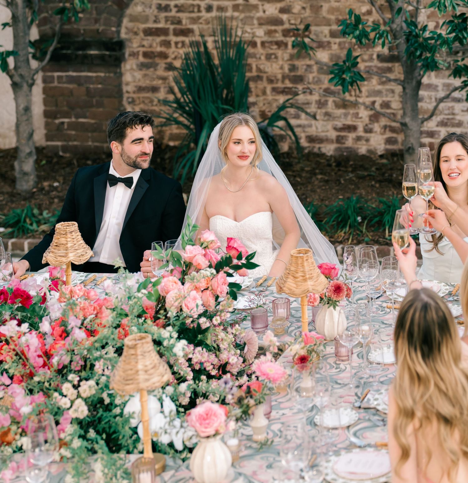 Long wedding reception table decorated with pink floral arrangements and glowing candles against brick walls.