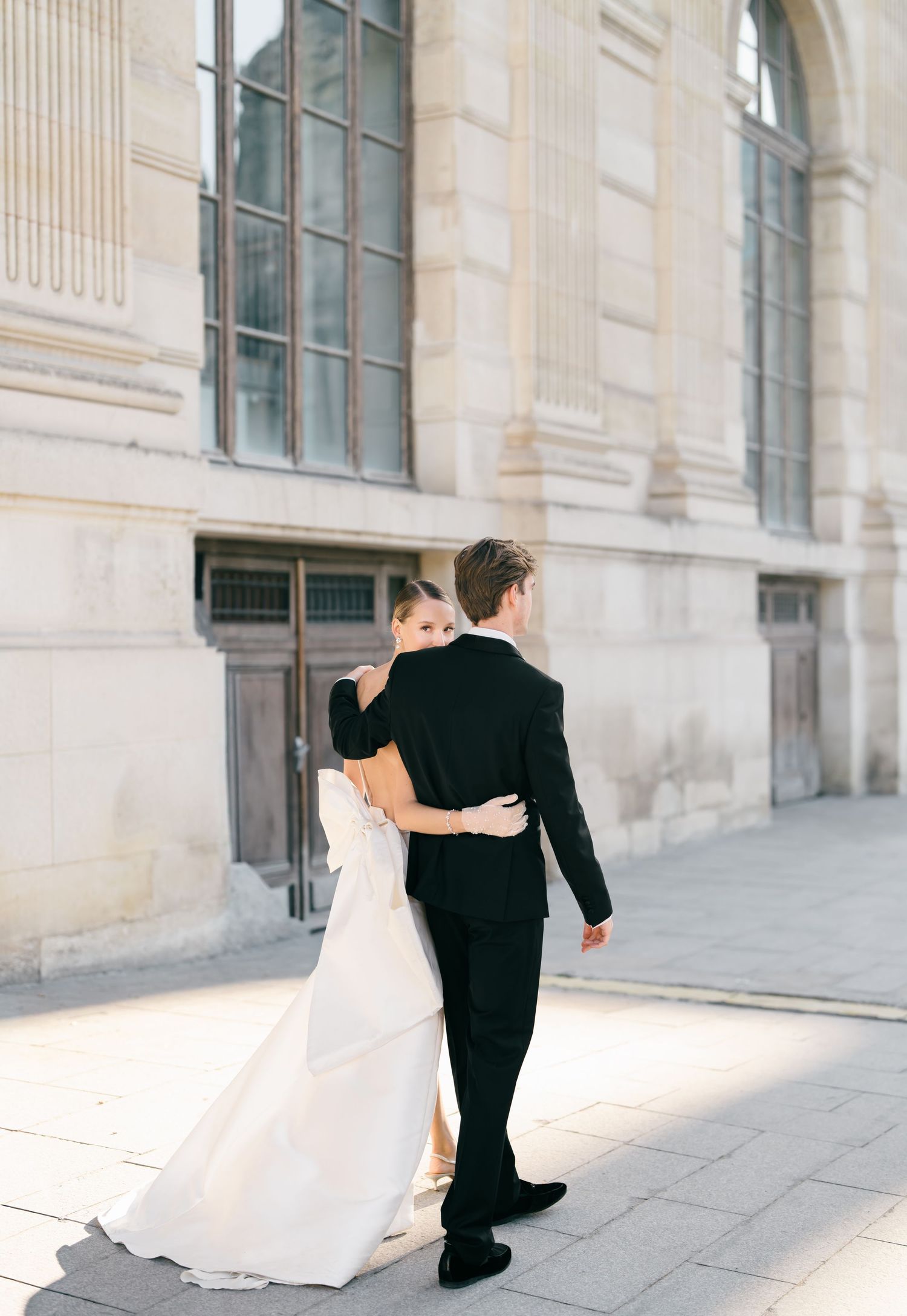 Bride and groom embrace in elegant wedding photos against a classic stone building with columns at The Louvre Museum. Paris, France.