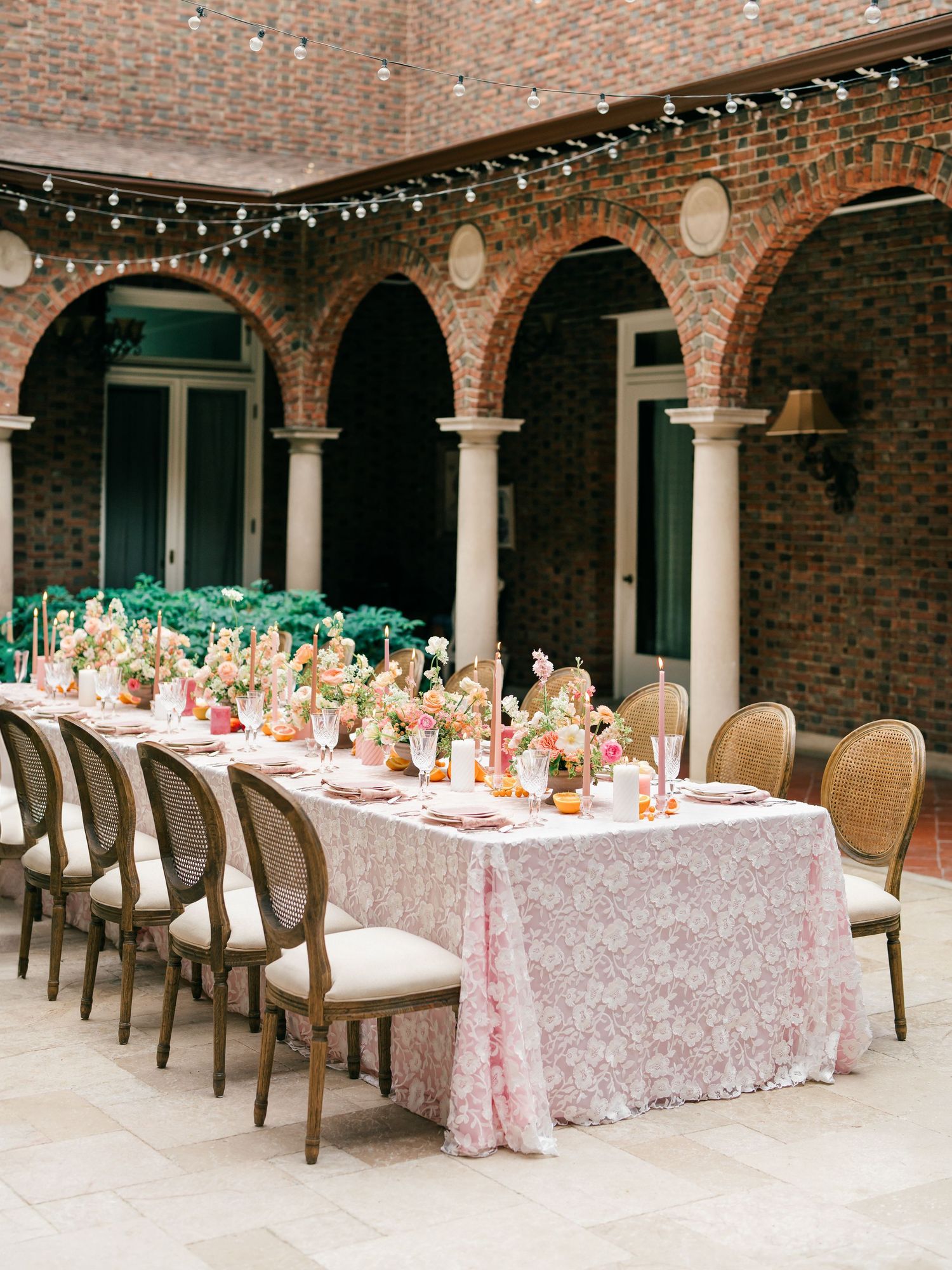Long banquet tables with pink tablecloths and floral centerpieces set under a rustic brick archway in an elegant courtyard.