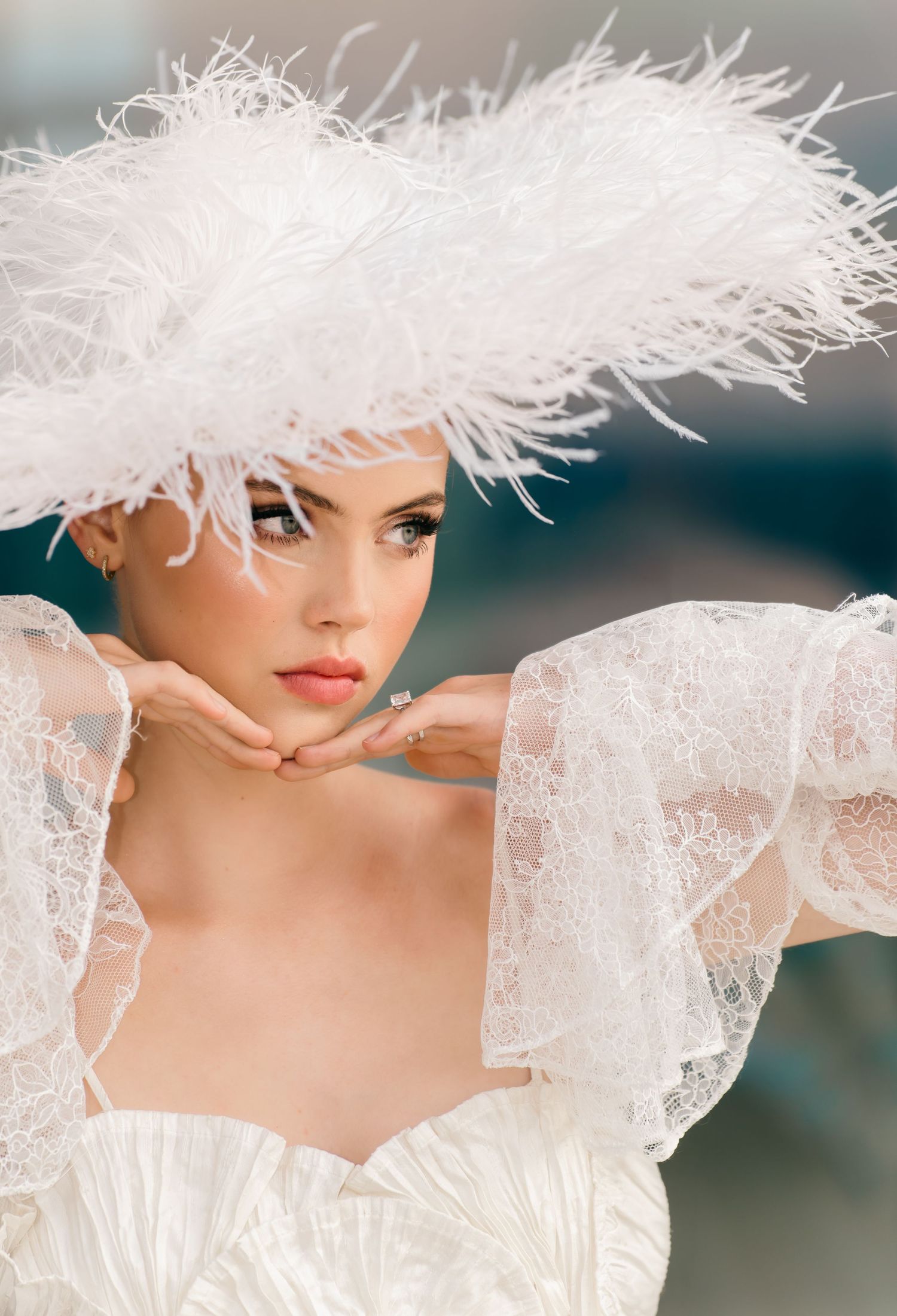 Elegant bride wearing a white feathered hat and lace gown, posing gracefully with hands near face.