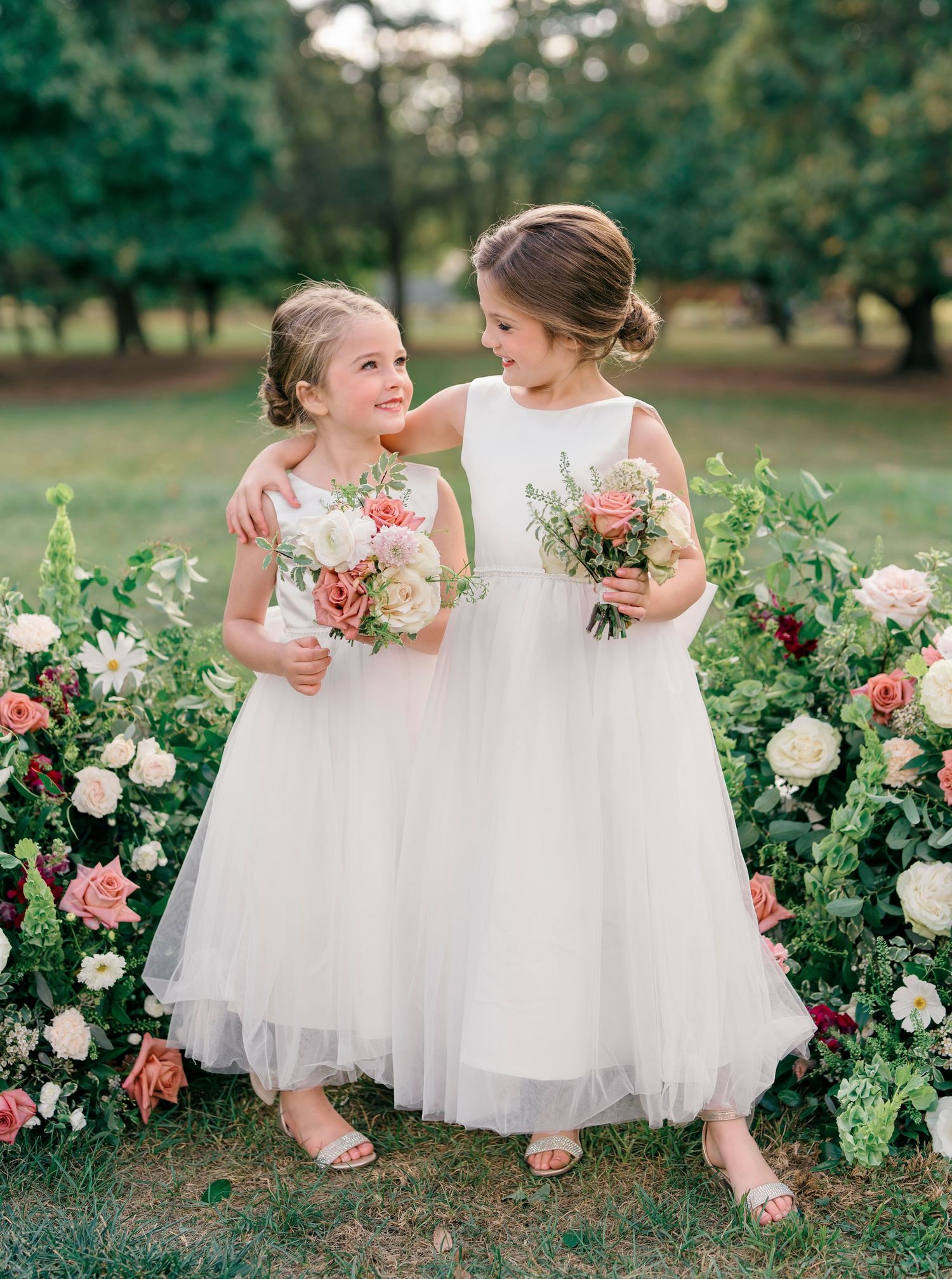 Two young attendants in white tulle dresses share a sweet moment among blooming garden roses during a wedding celebration.