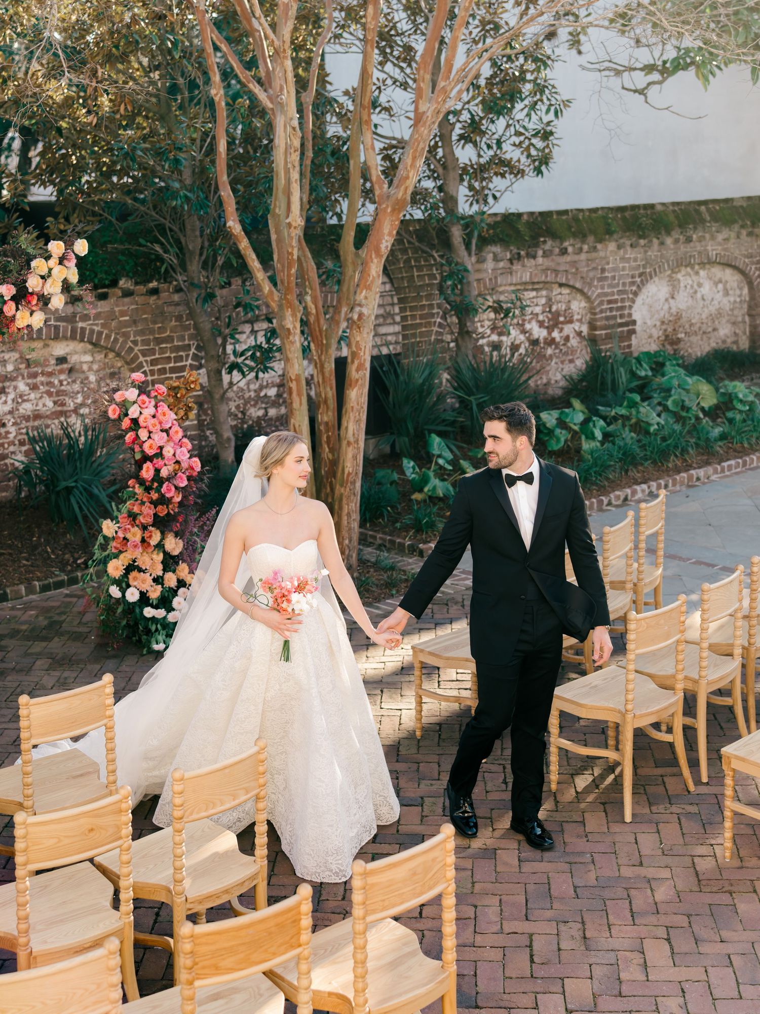 Romantic outdoor wedding ceremony setup with wooden chairs and floral arrangements on a brick patio at sunset.
