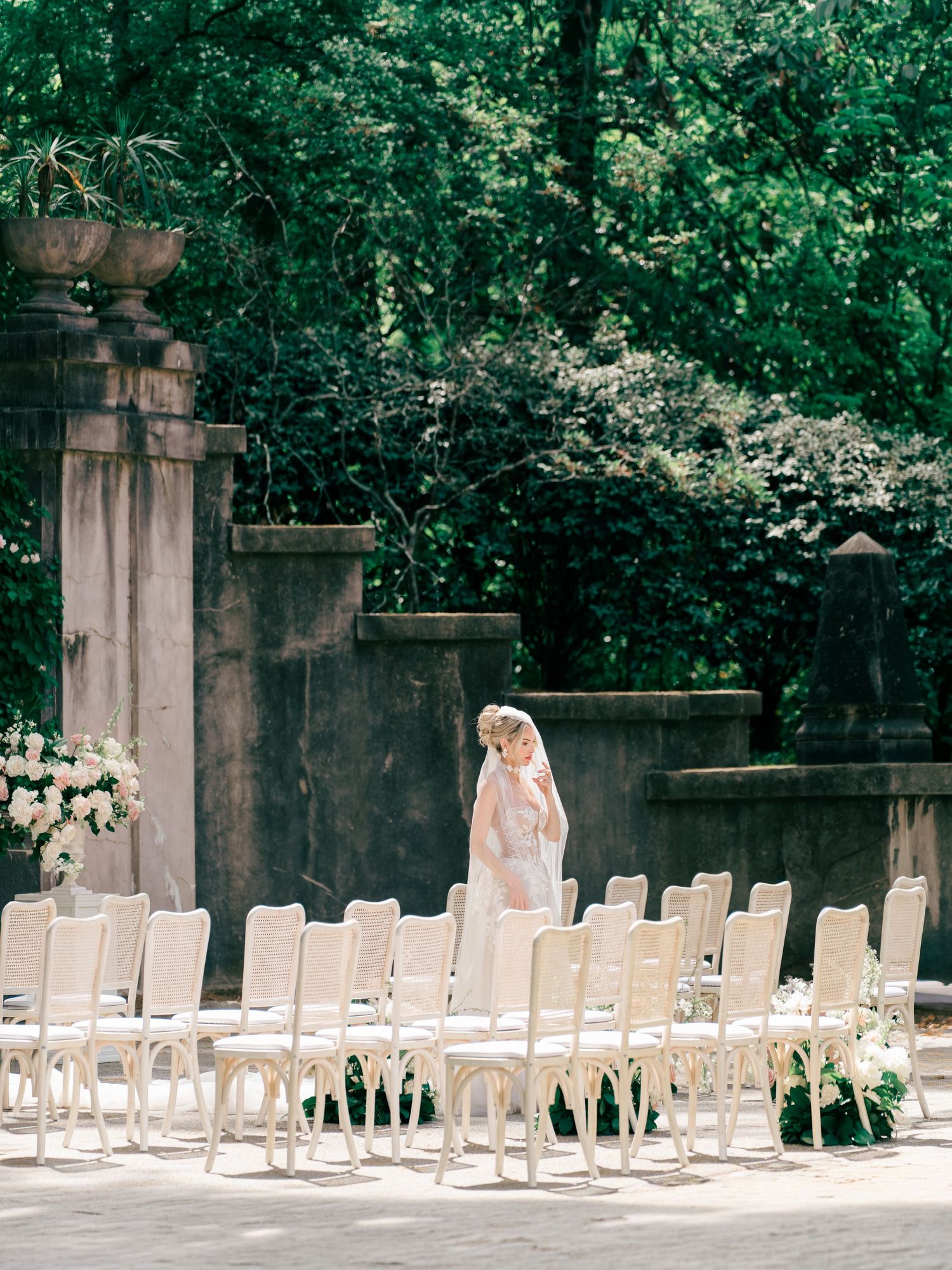 Bride standing among white chairs at an elegant outdoor wedding ceremony setup surrounded by lush greenery.
