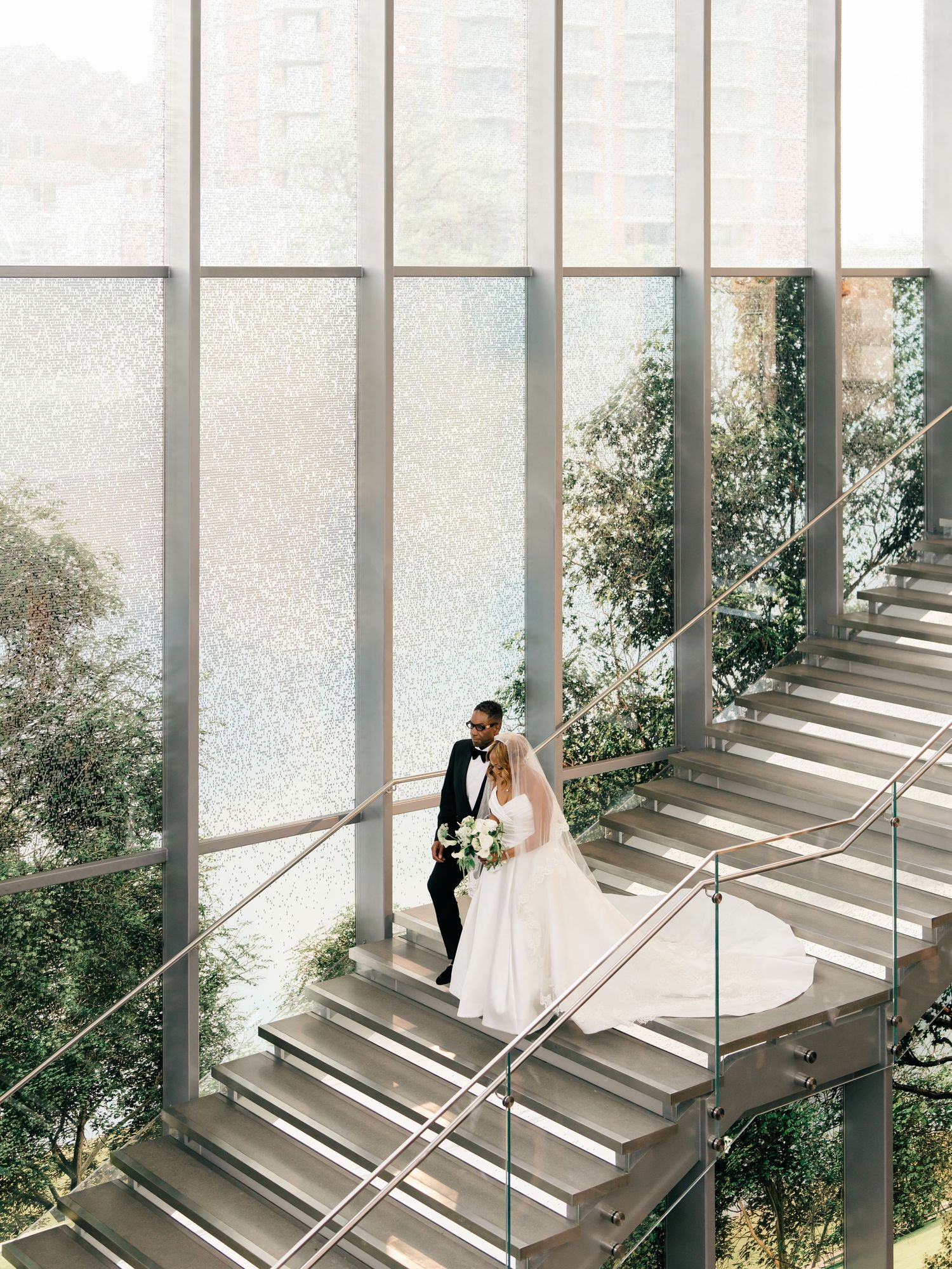 Modern interior staircase with glass walls and natural light showcases a couple in wedding attire ascending the steps.