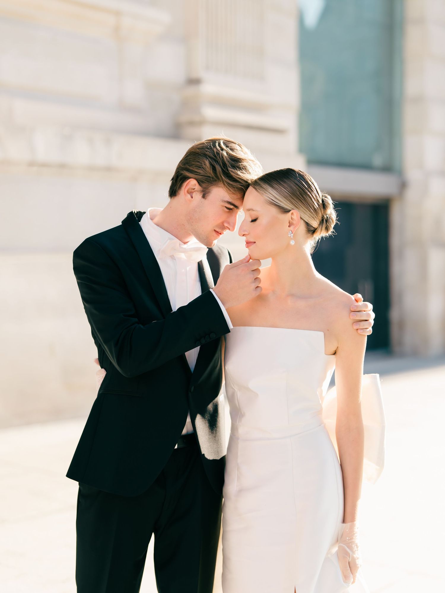 A romantic wedding couple, in formal attire, share an intimate moment against a light architectural backdrop in Paris, France.