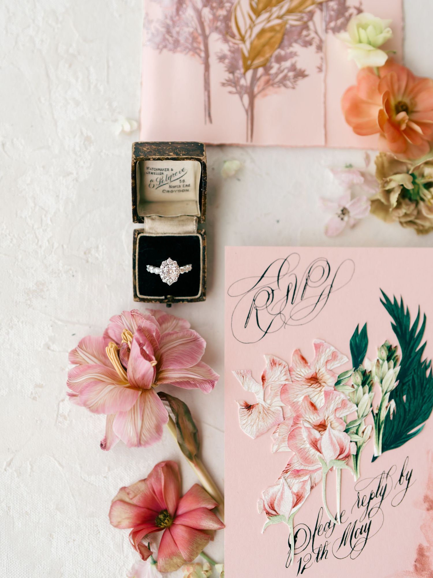 Elegant engagement ring in vintage box with pink floral wedding RSVP stationery and fresh flowers flatlay.