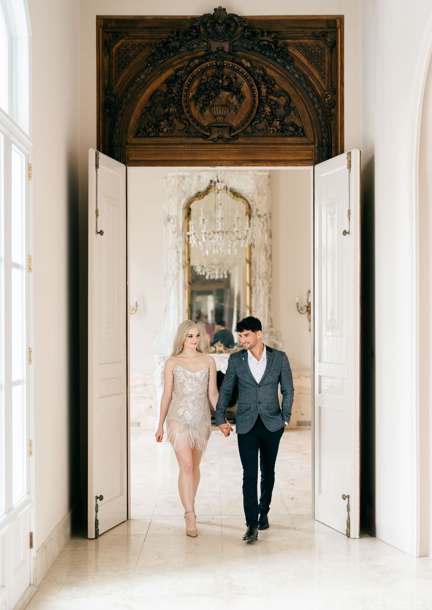 Bride and groom stroll down bright white corridor with ornate architectural details and natural lighting.