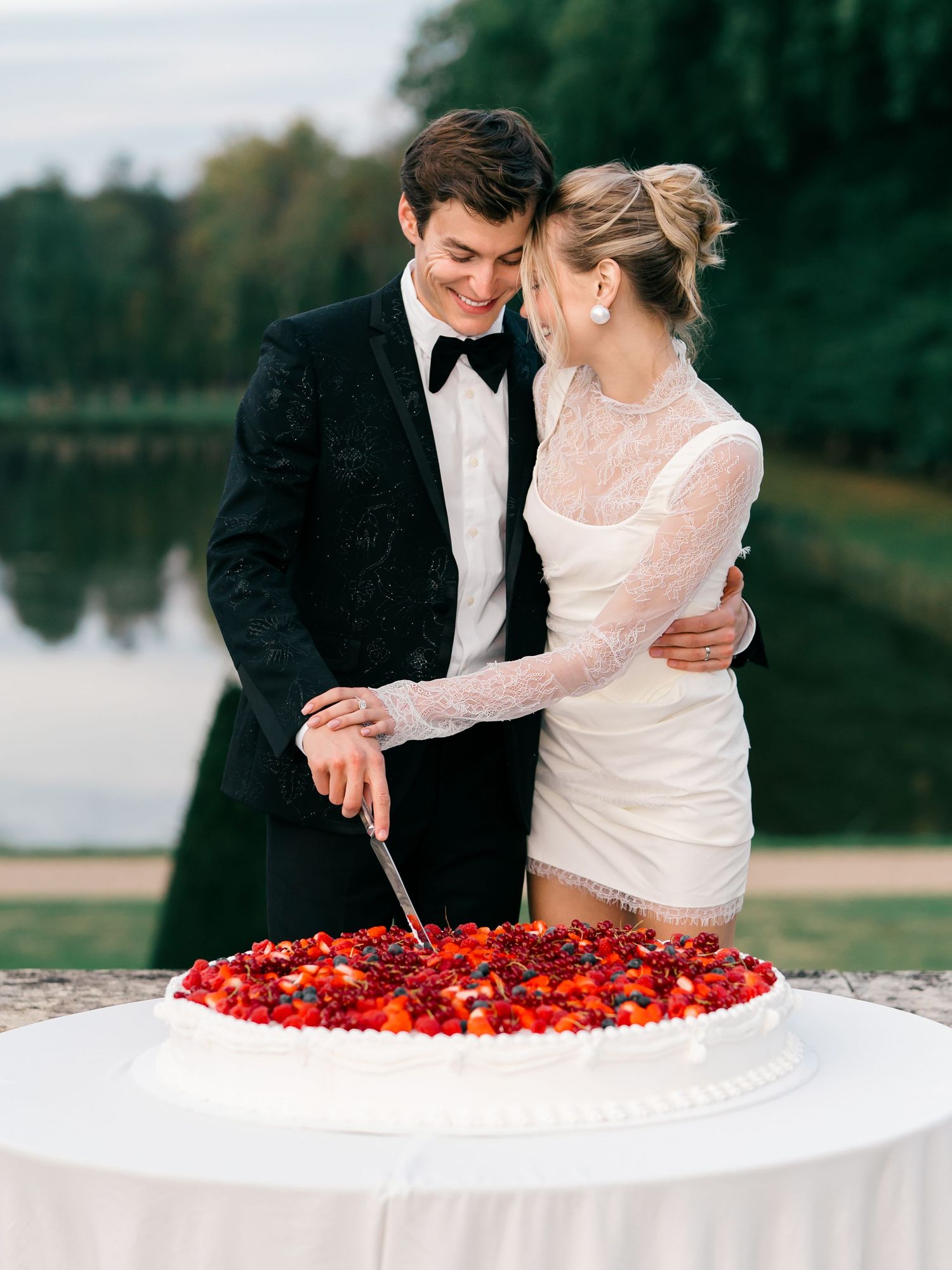 A couple cuts into a large round wedding cake decorated with fresh berries. Outdoor reception at Chateau de Villette in Paris, France.