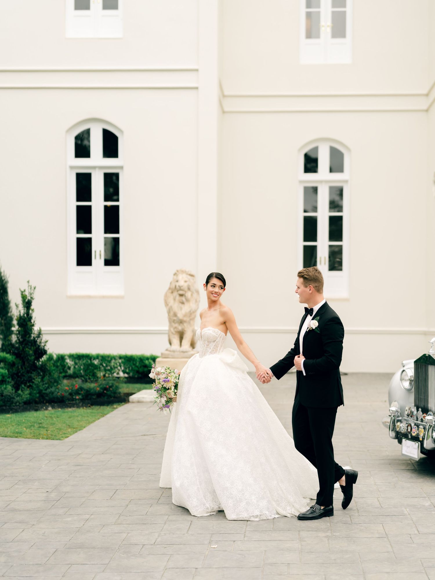 A romantic wedding couple walking together outside a classic white building with architectural details.