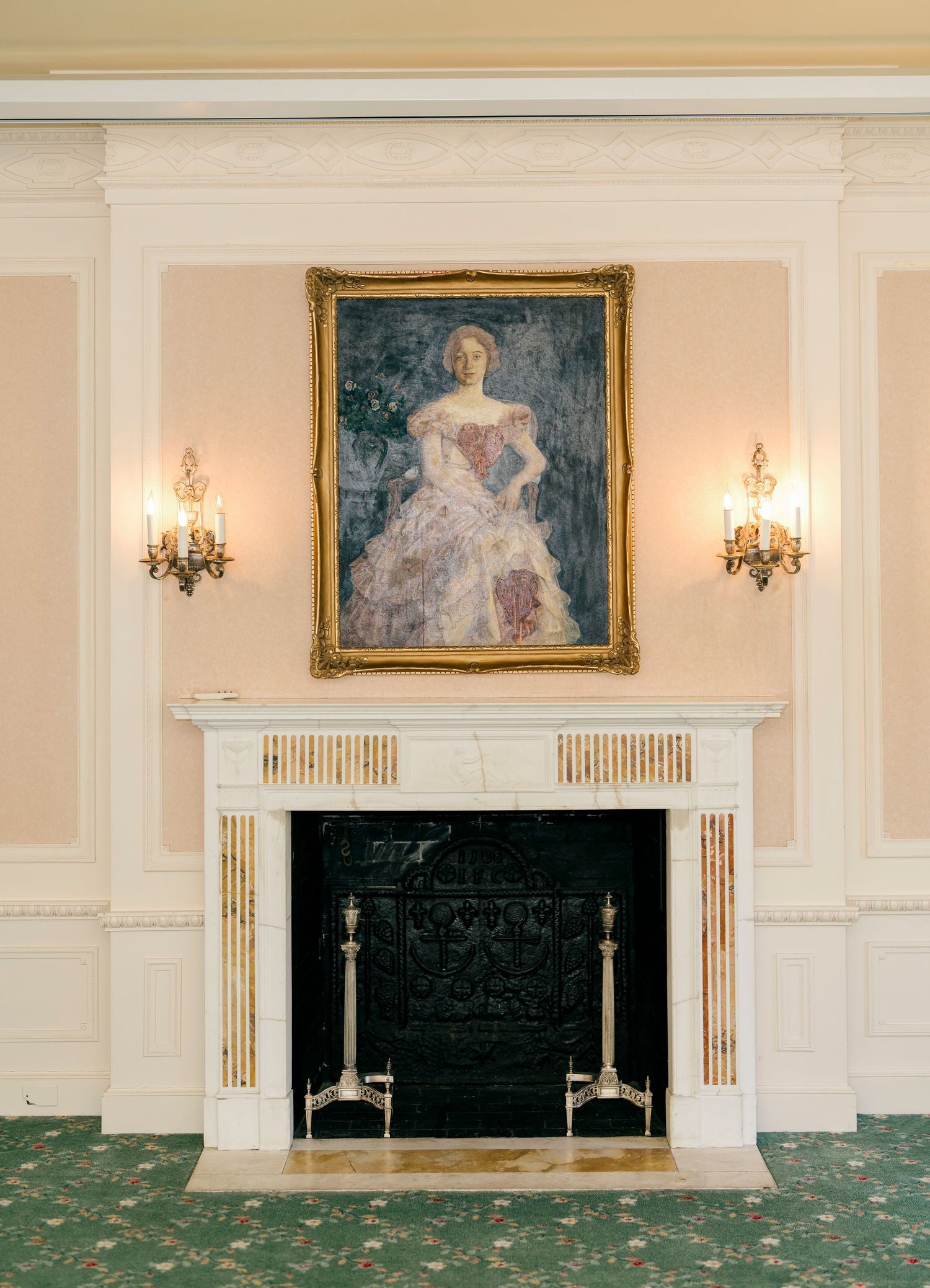 Elegant white fireplace with ornate gold-framed portrait and wall sconces in a formal room with green carpeting.
