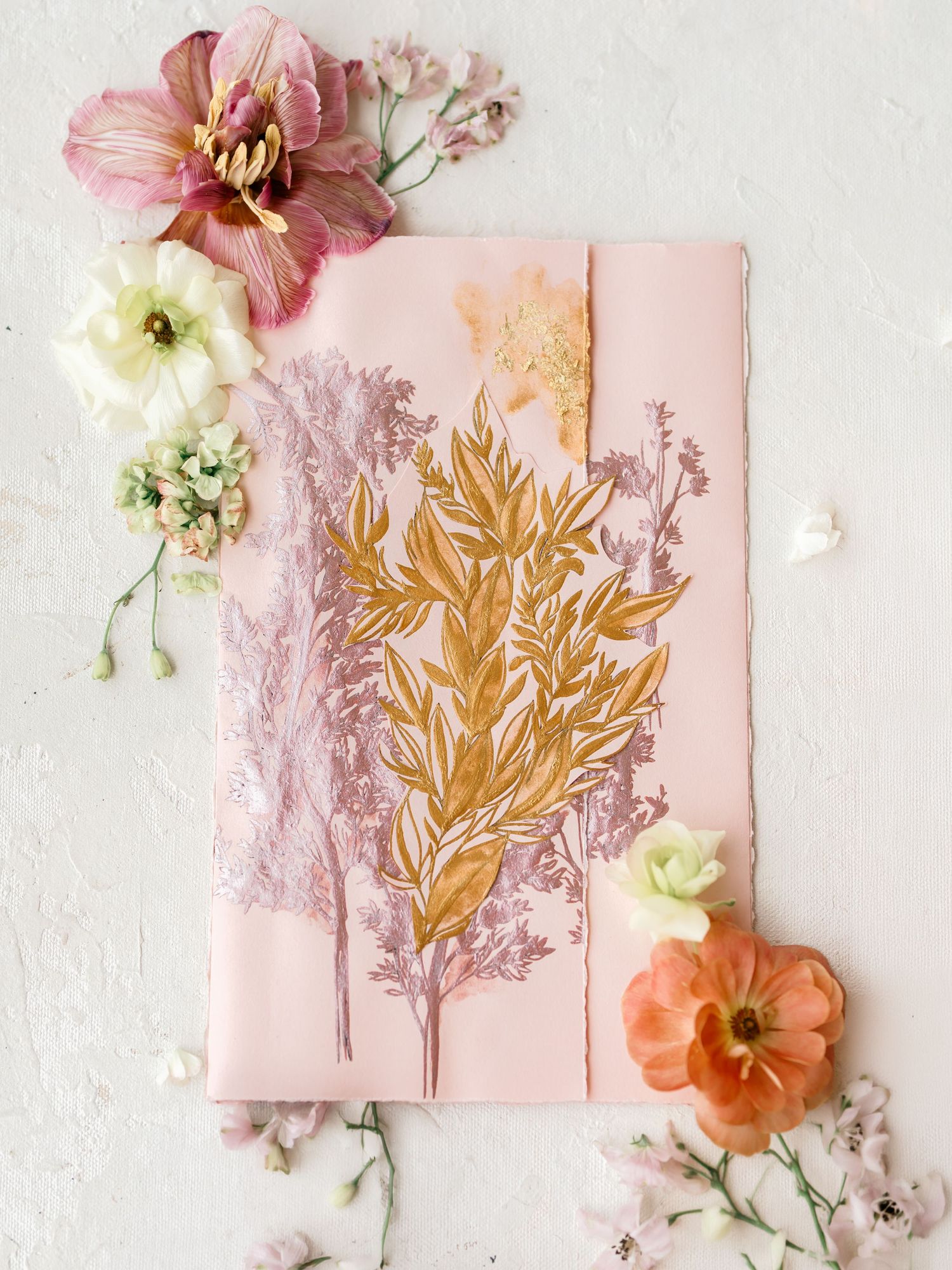 Elegant pink wedding stationery with gold botanical prints surrounded by soft pink and white flowers.