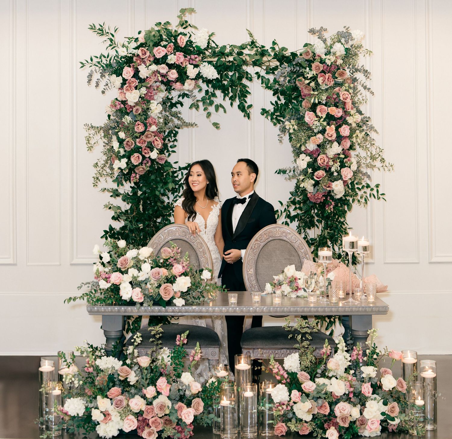 Couple stands at floral wedding arch with pink roses and greenery, elegant sweetheart table with candles below.