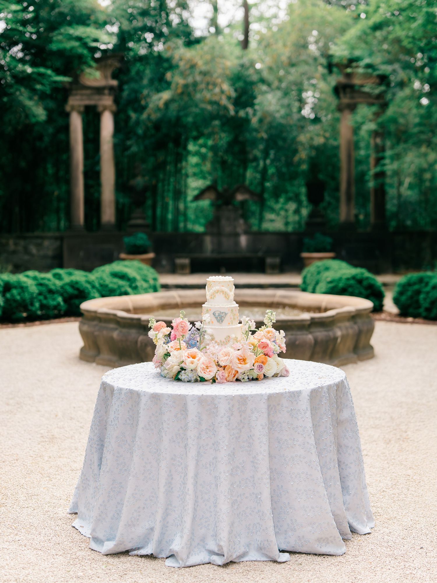 Elegant wedding cake with pink florals on a silver tablecloth in a garden with stone fountain and columns.