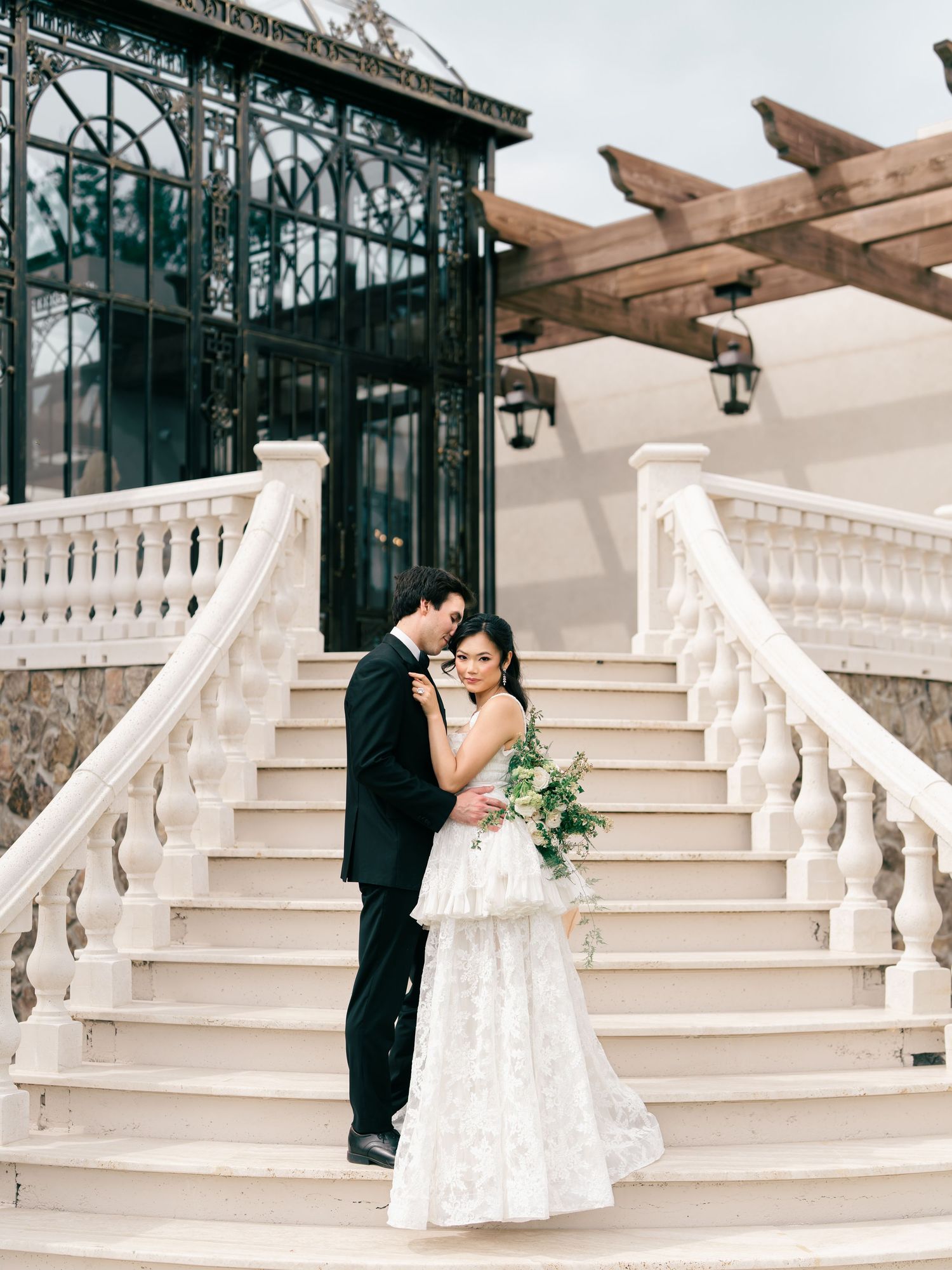Couple embraces on grand white staircase, groom in black tuxedo, bride holding greenery bouquet.