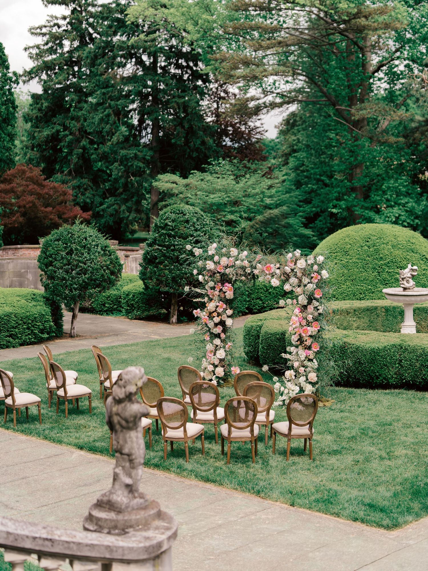 Elegant garden wedding ceremony setup with vintage chairs and floral arch arrangements in a lush green outdoor setting.
