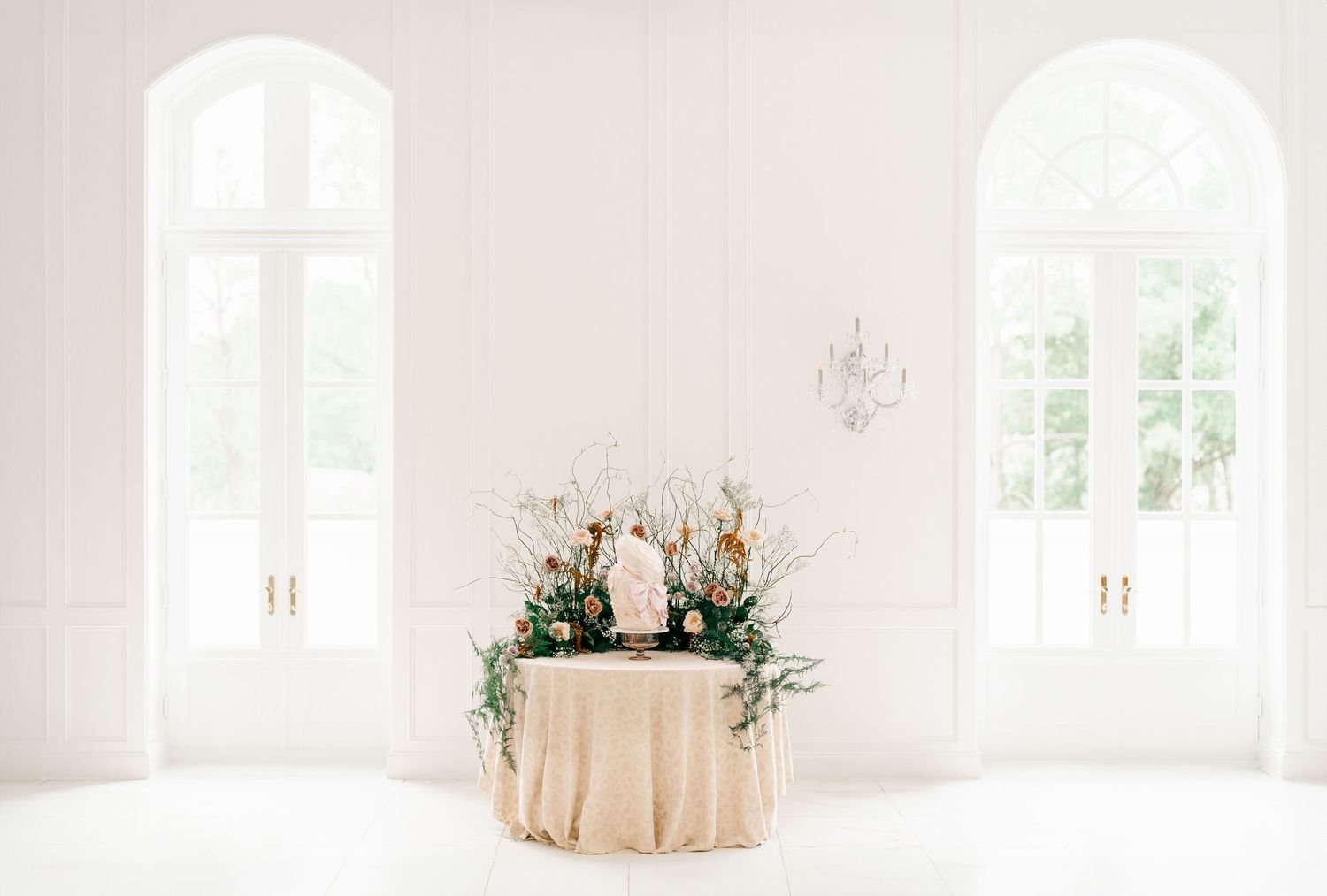 Elegant wedding table setup with floral centerpiece in bright white room with arched windows and natural light.