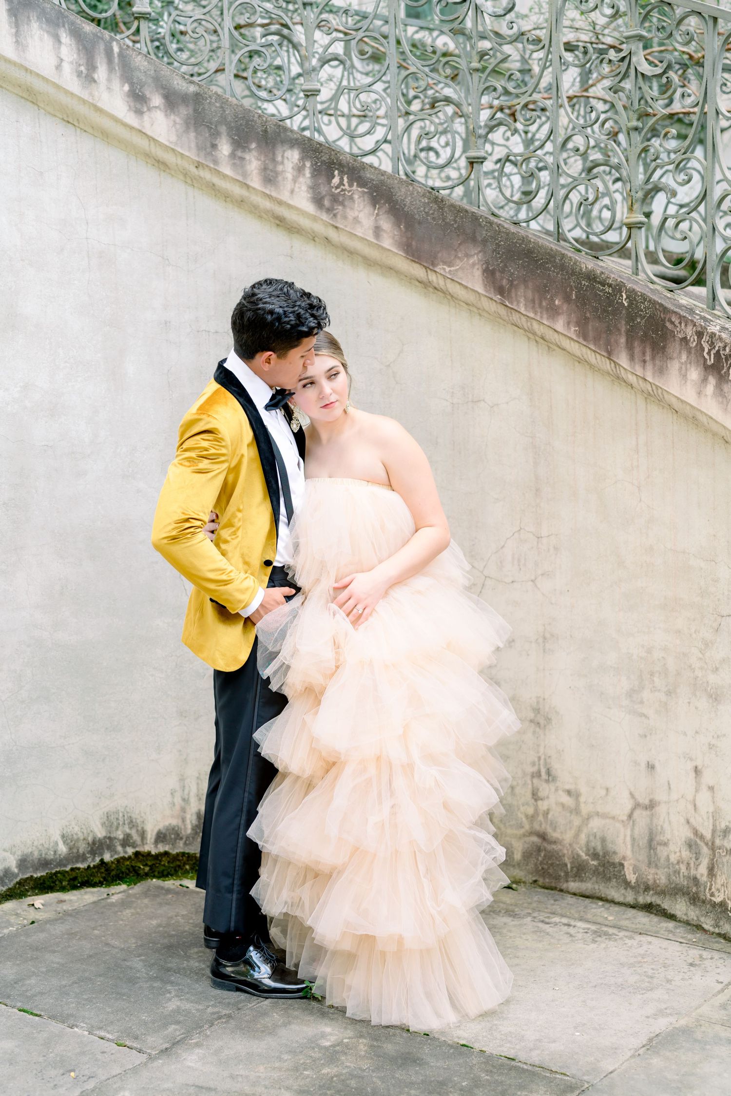 Romantic couple in peach tulle gown and yellow suit jacket against ornate wall.