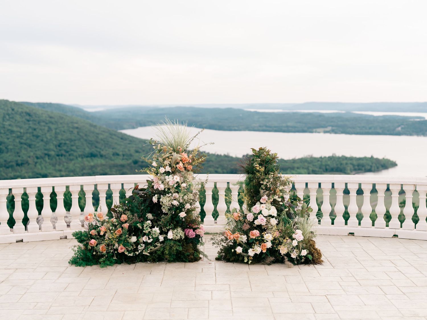 Elegant outdoor wedding ceremony setup with lush floral arrangements on a terrace overlooking a scenic lake and green hills.