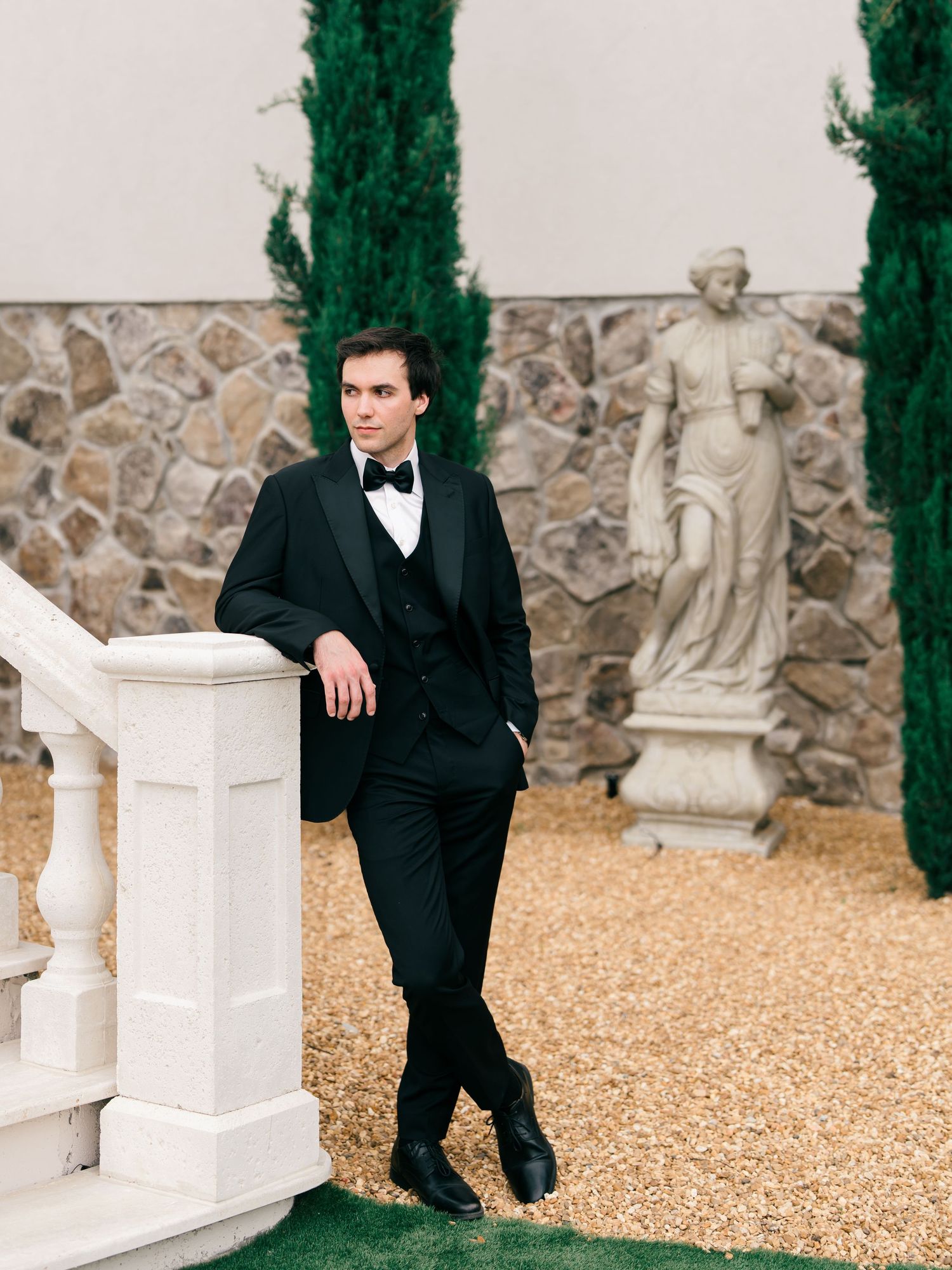 Groom in black tuxedo with bow tie leaning against white balustrade near stone wall and classical statue.