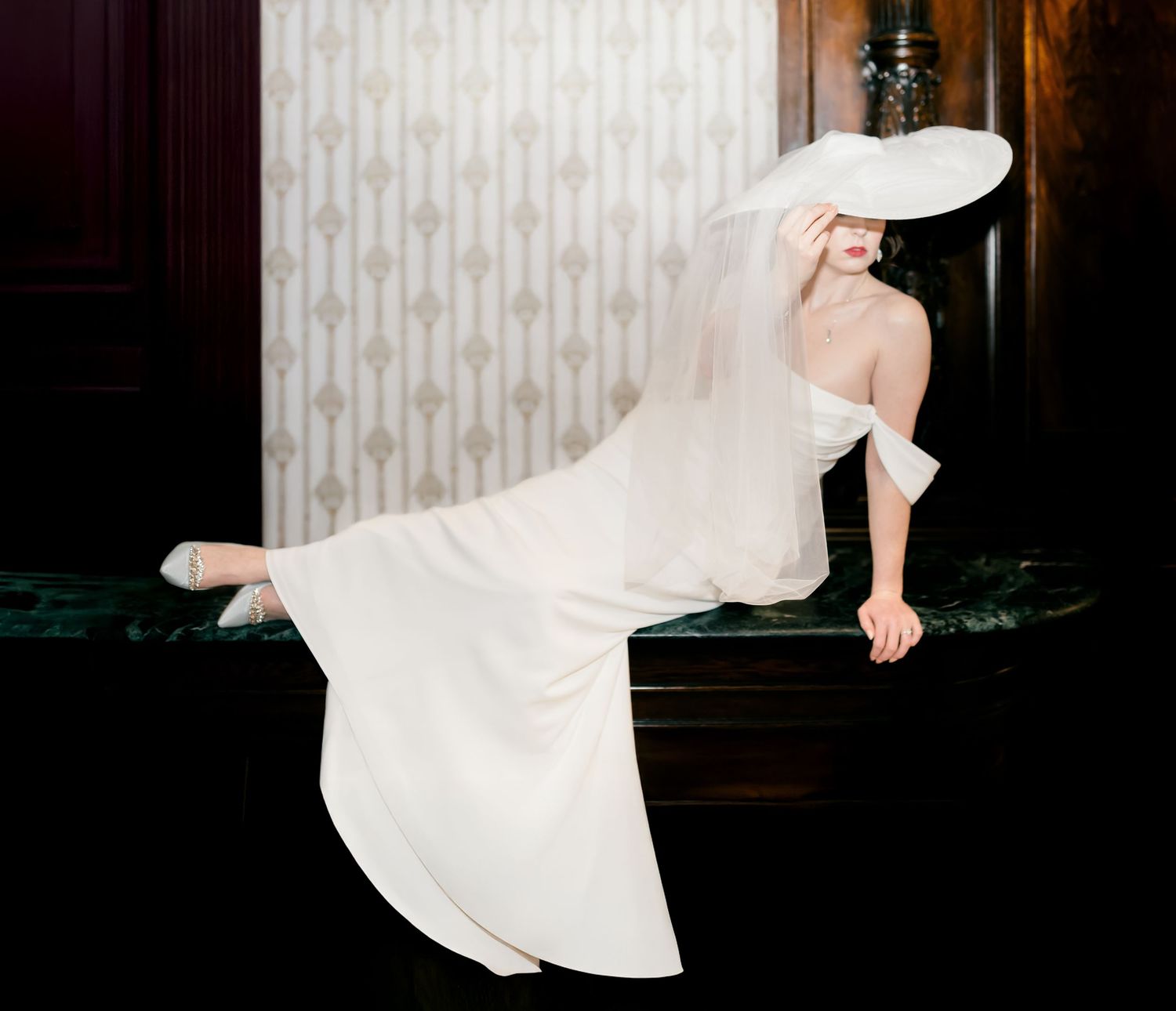 A figure in a flowing white wedding dress and hat strikes an elegant pose against a vintage wallpapered wall.