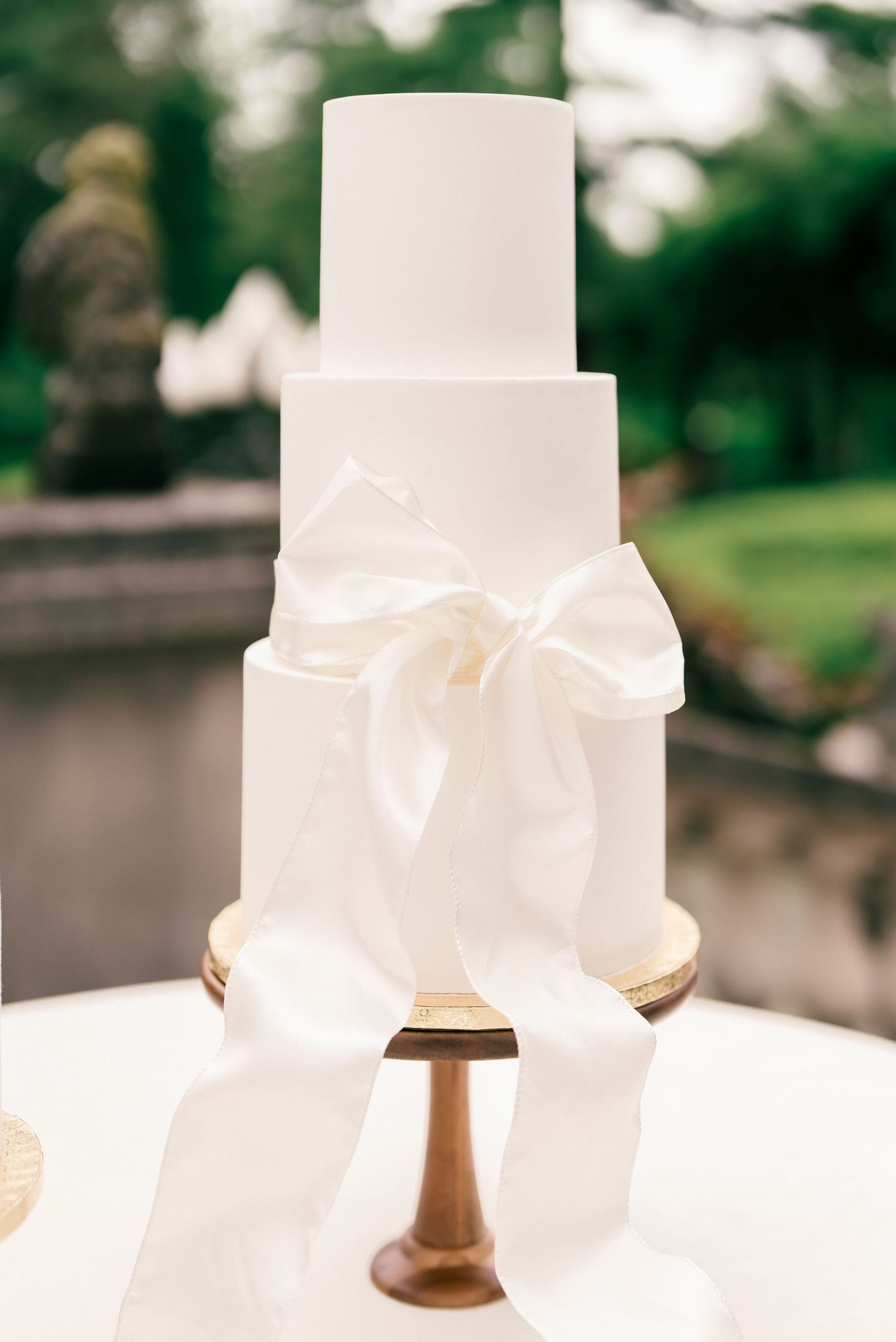 A series of elegant white wedding cakes with ribbon details displayed on wooden cake stands against a blurred outdoor background.