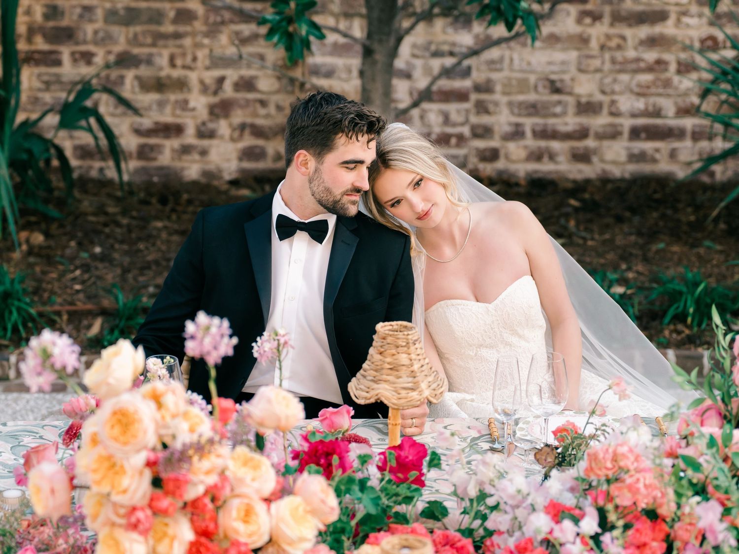 Couple seated at elegant wedding reception table with vibrant coral and pink floral centerpieces against rustic brick wall.