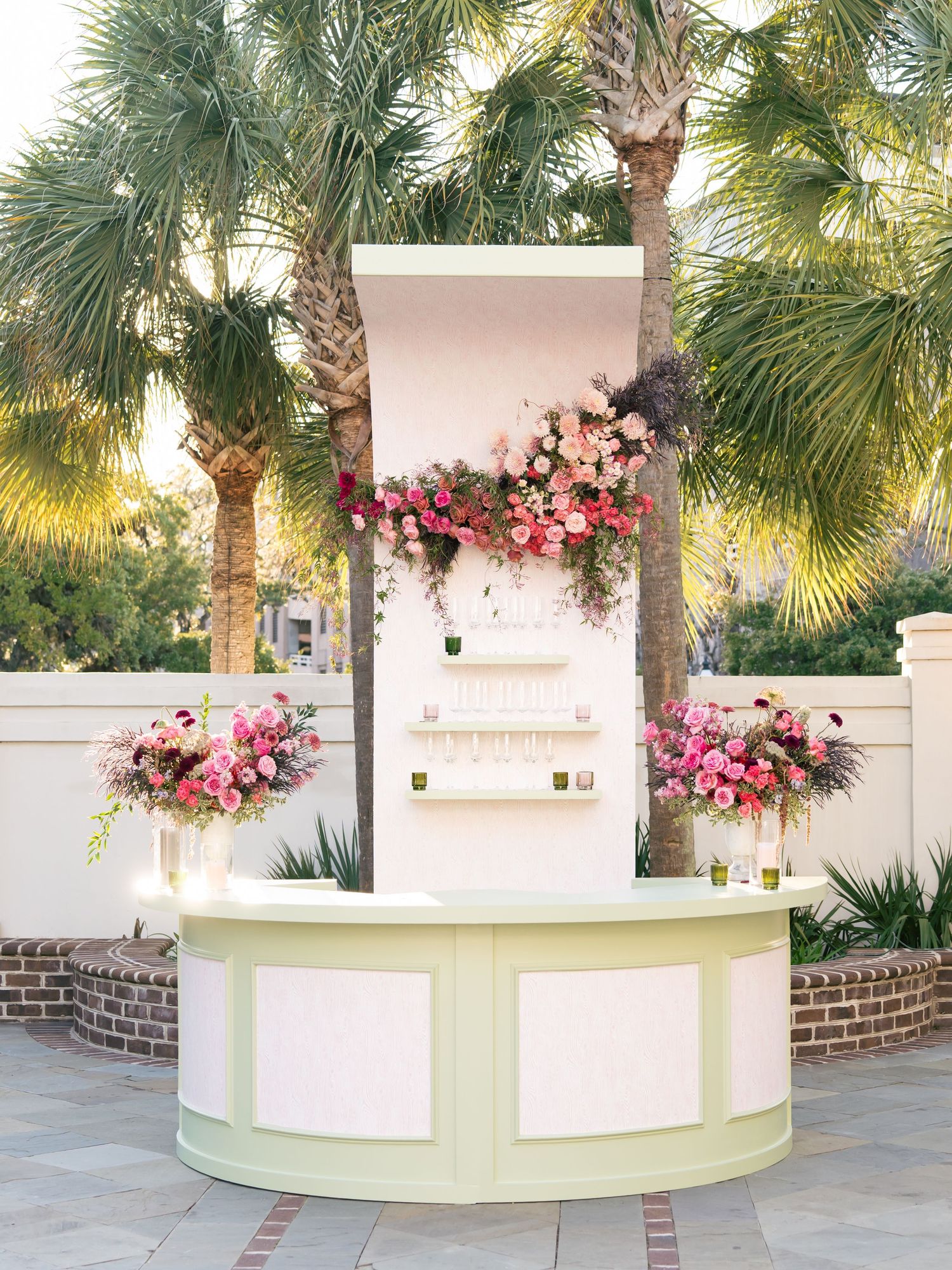 Curved white bar counter decorated with pink and red floral arrangements under palm trees at an outdoor venue.