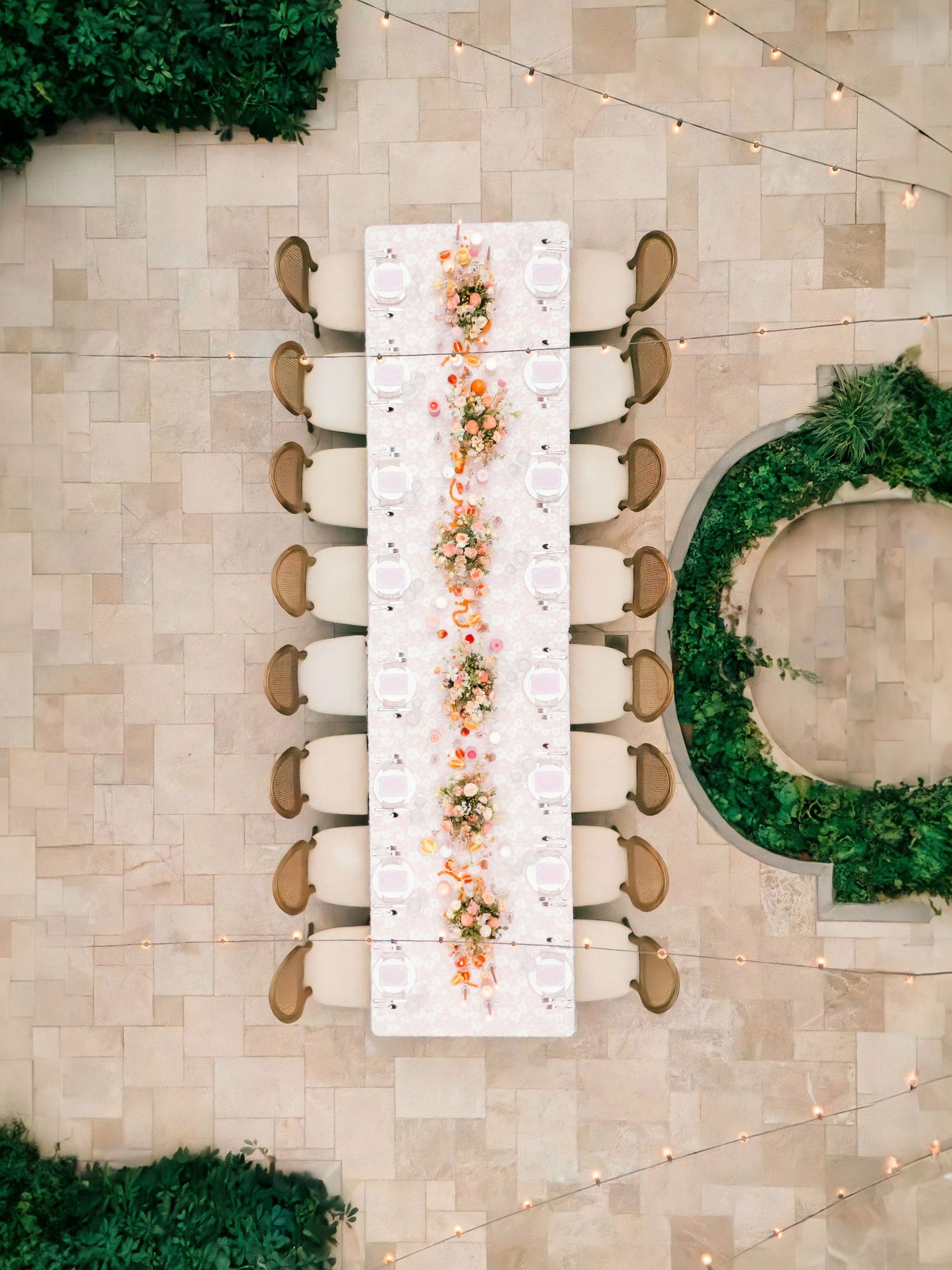 Aerial view of a long white dining table with chairs on a stone patio surrounded by lush greenery and string lights.