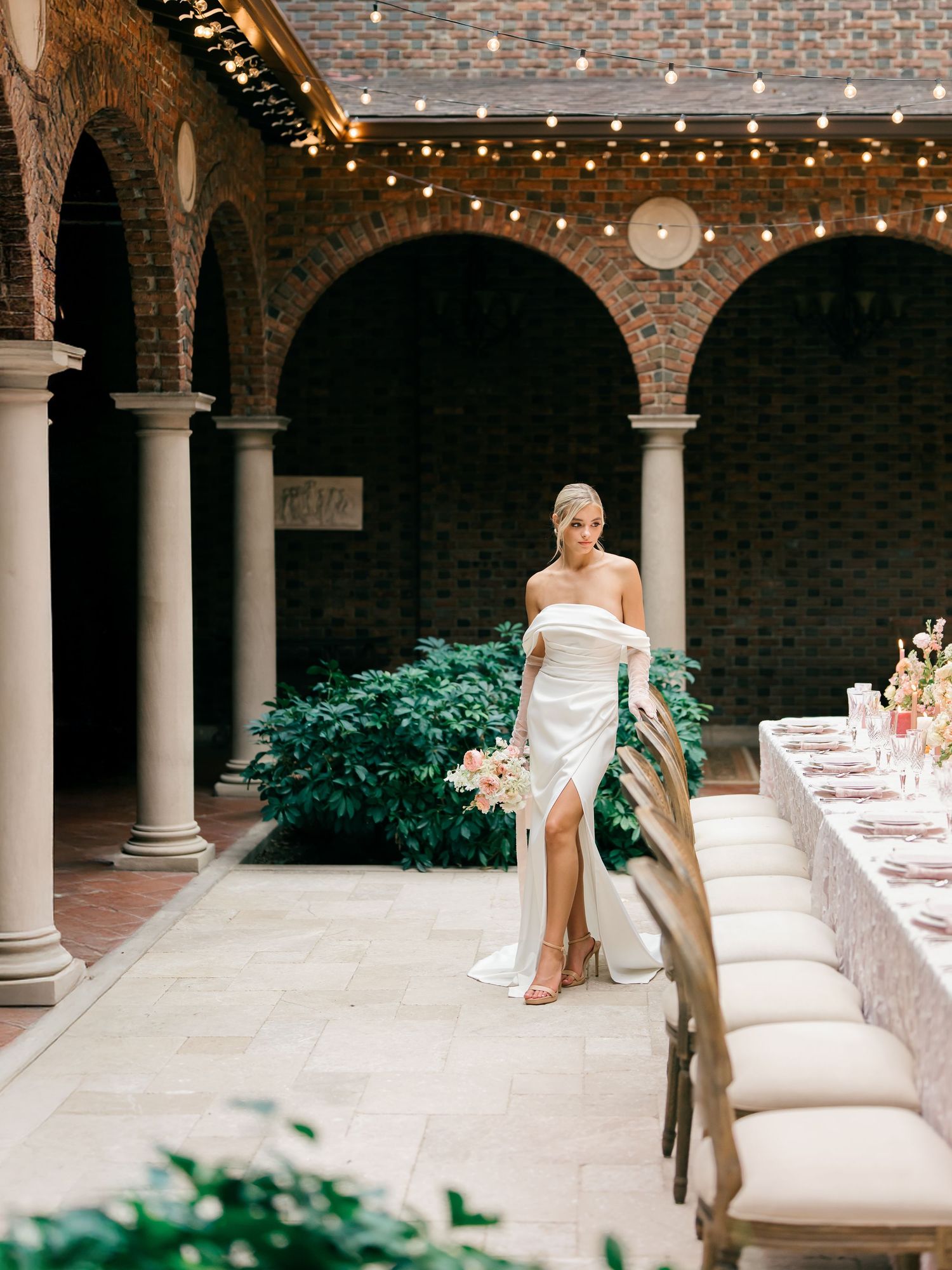 A bride in white dress sits elegantly on a stone bench in a courtyard with brick walls and classic columns.