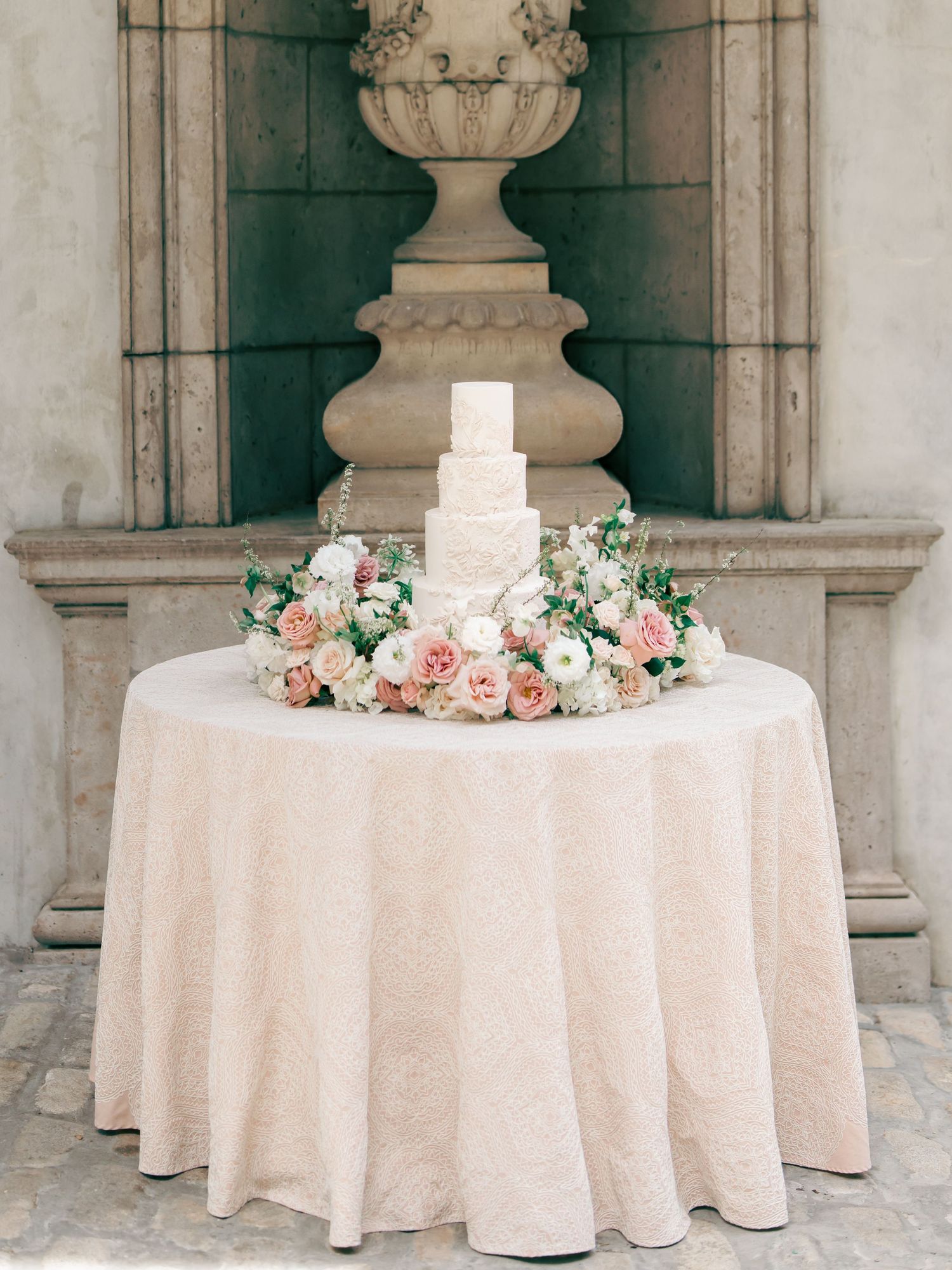 Elegant white tiered wedding cake surrounded by blush pink and white roses on a lace-covered round table.