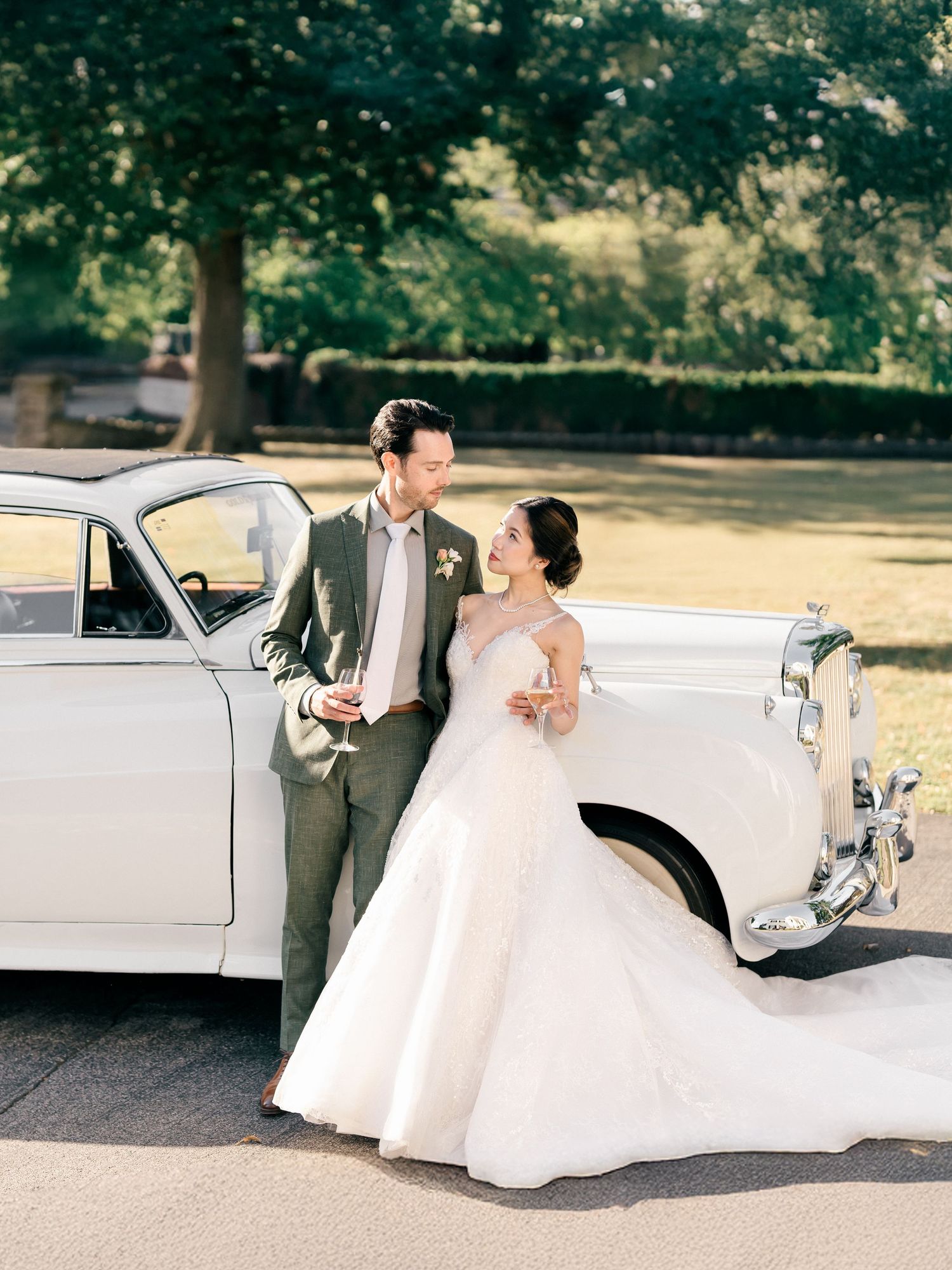 A couple poses next to a vintage white classic car wearing elegant wedding attire in a lush outdoor setting.