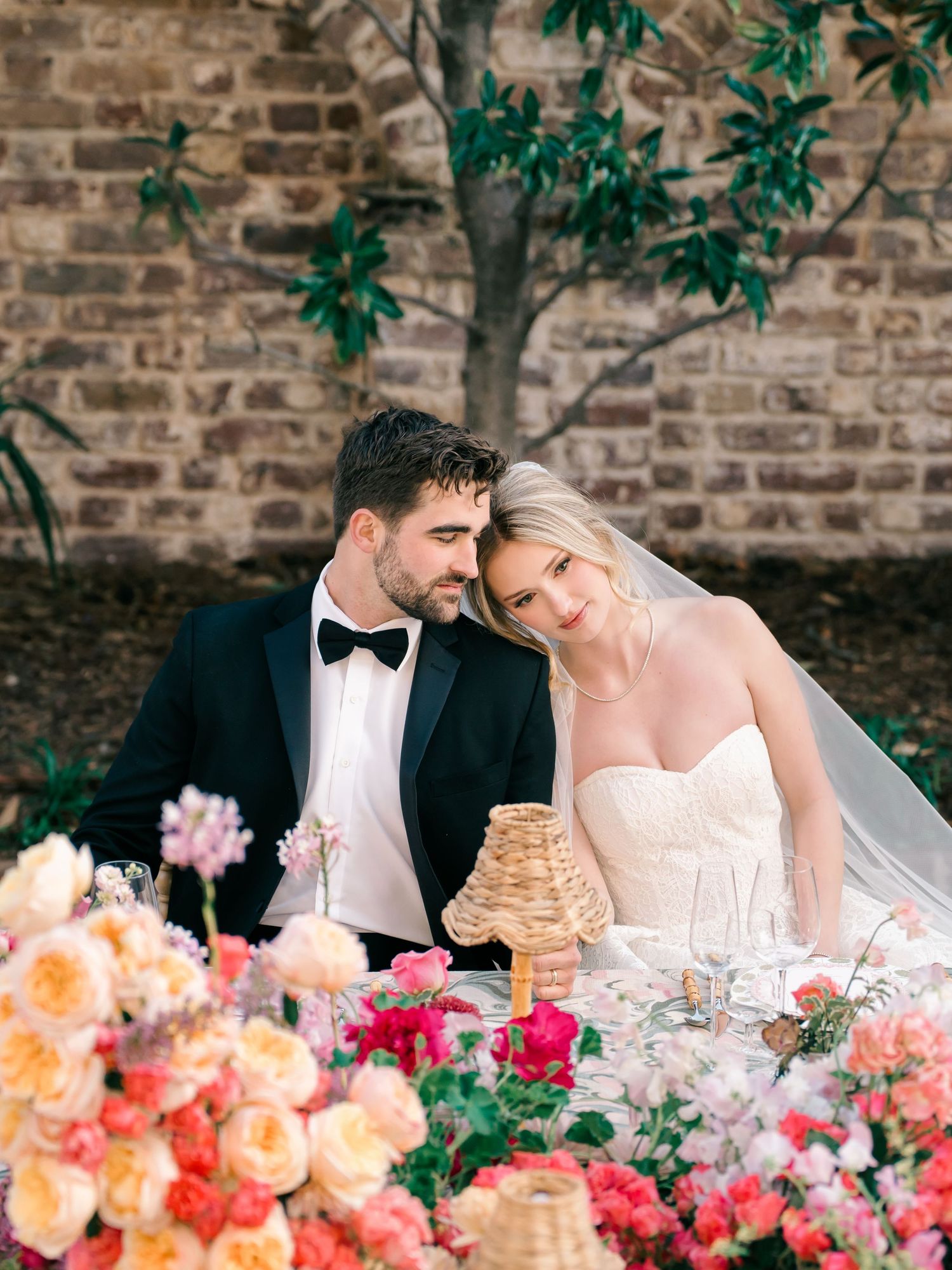 A romantic wedding couple sits at an elegantly decorated table with vibrant coral and pink floral arrangements against a brick wall.
