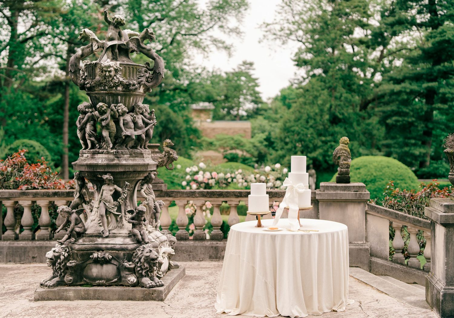 Elegant wedding cake table setup on a stone terrace with ornate vintage sculpture and lush garden backdrop.
