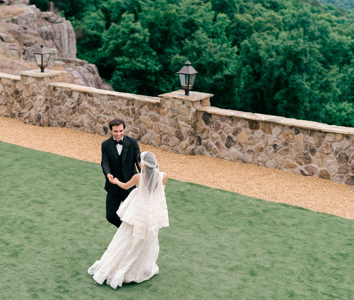 Bride in white gown and groom in black tux share first look on green lawn by stone wall.
