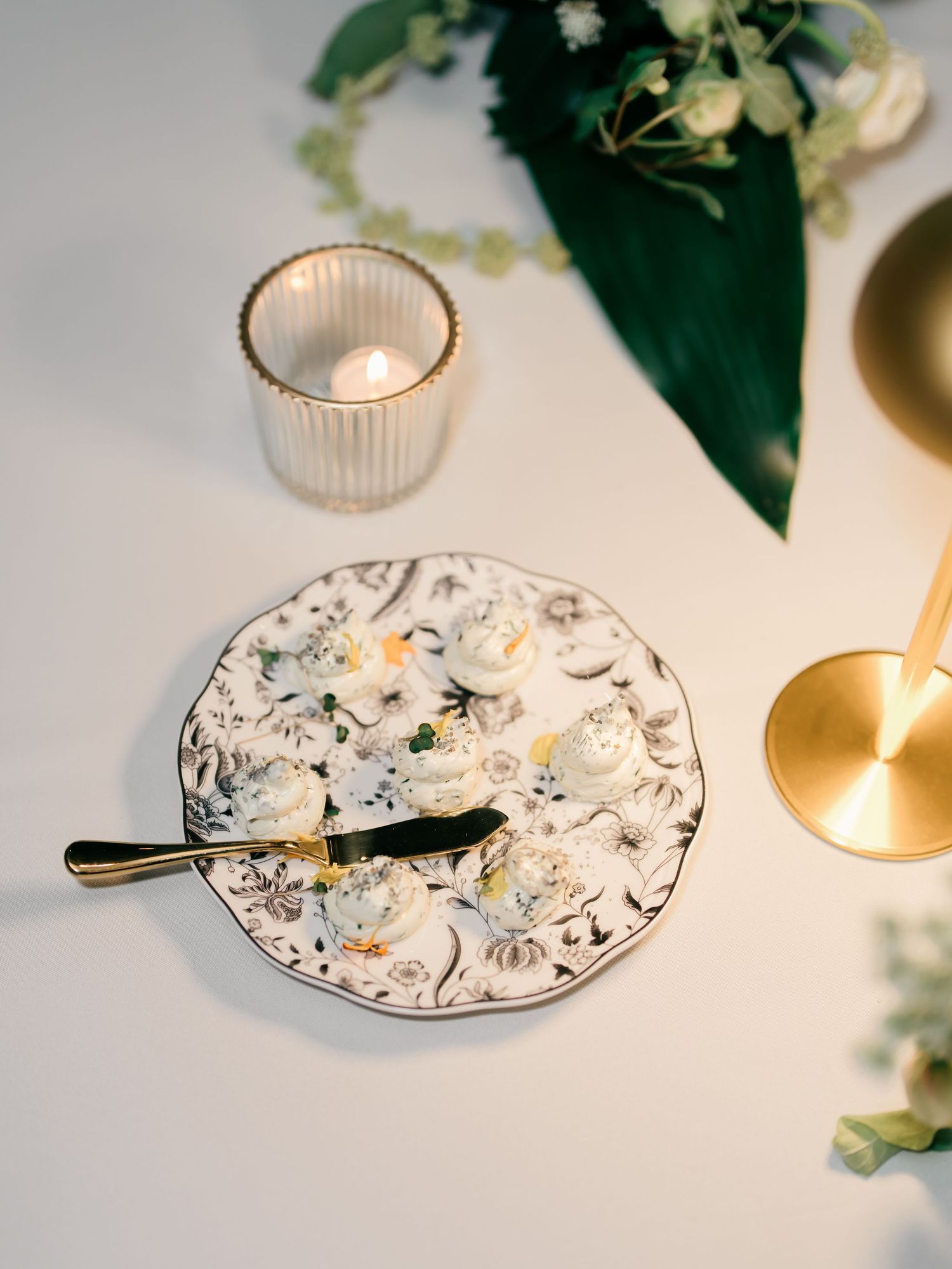 Elegant table setting with floral patterned plate, gold cutlery, and glowing candle votive against white background.